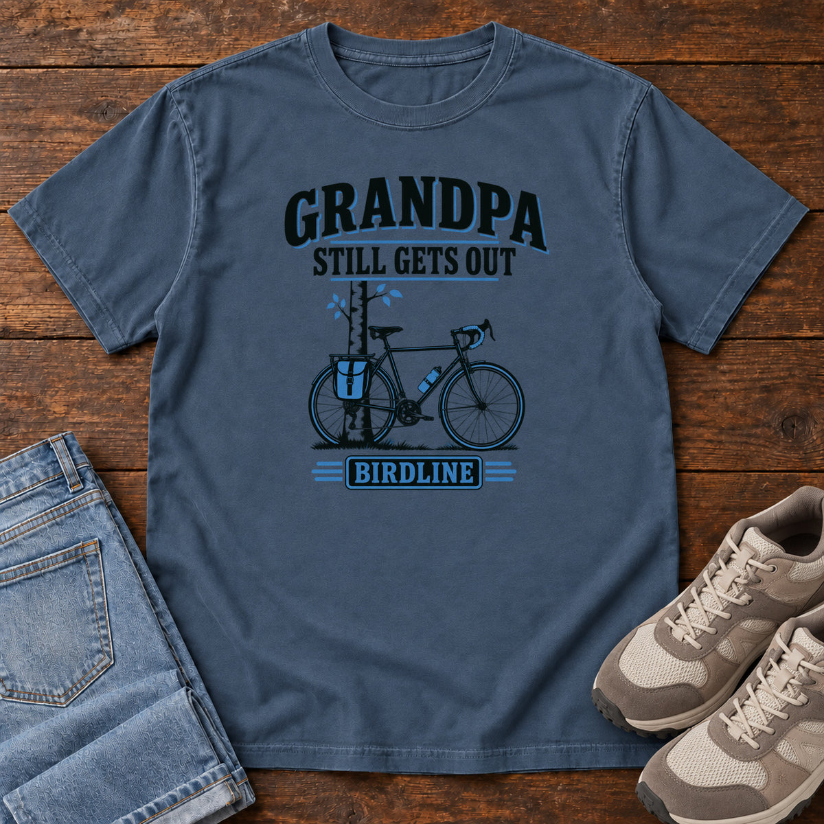 Midnight Grandpa Still Gets Out T-Shirt with a bicycle beside a slender tree and blue-accented lettering, front flat lay on wood.