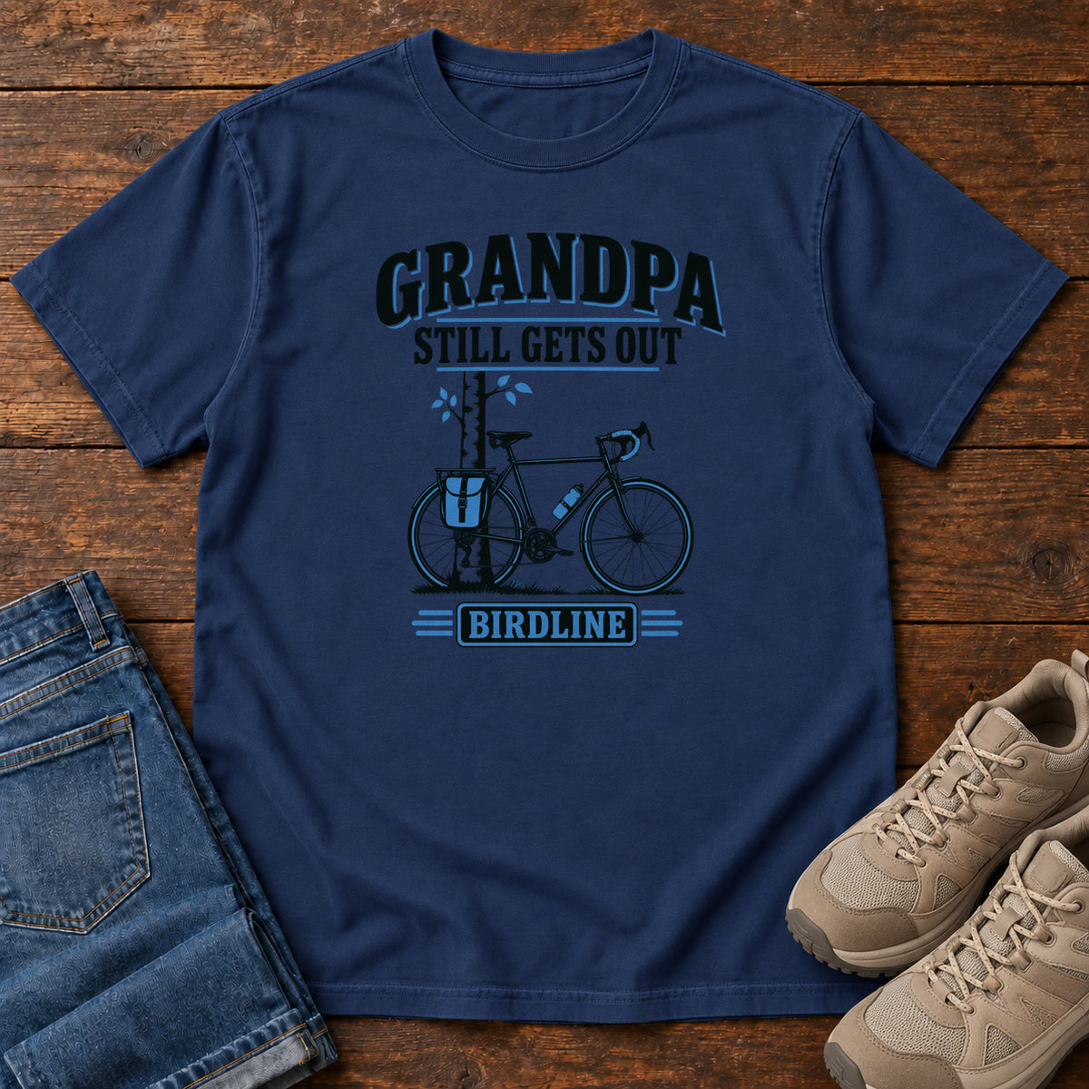 True Navy Grandpa Still Gets Out T-Shirt with a bicycle beside a slender tree and blue-accented lettering, front flat lay on wood.