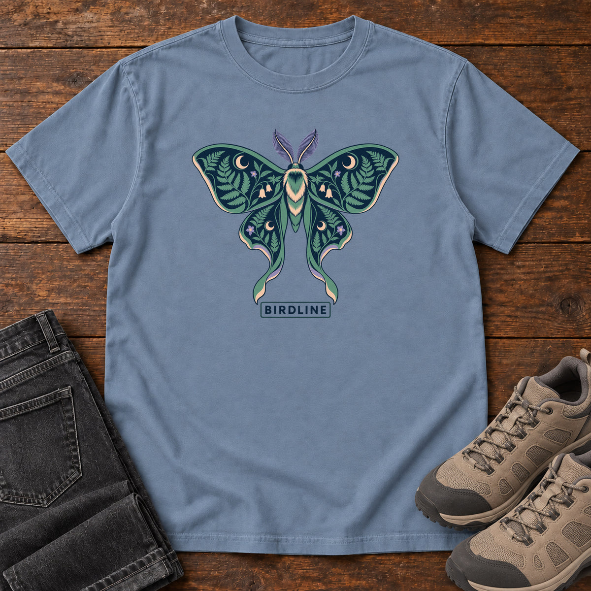 Botanical Moth T-Shirt in Blue Jean, front laid flat on wood, featuring a teal moth with patterned wings, leaves and crescent moons.