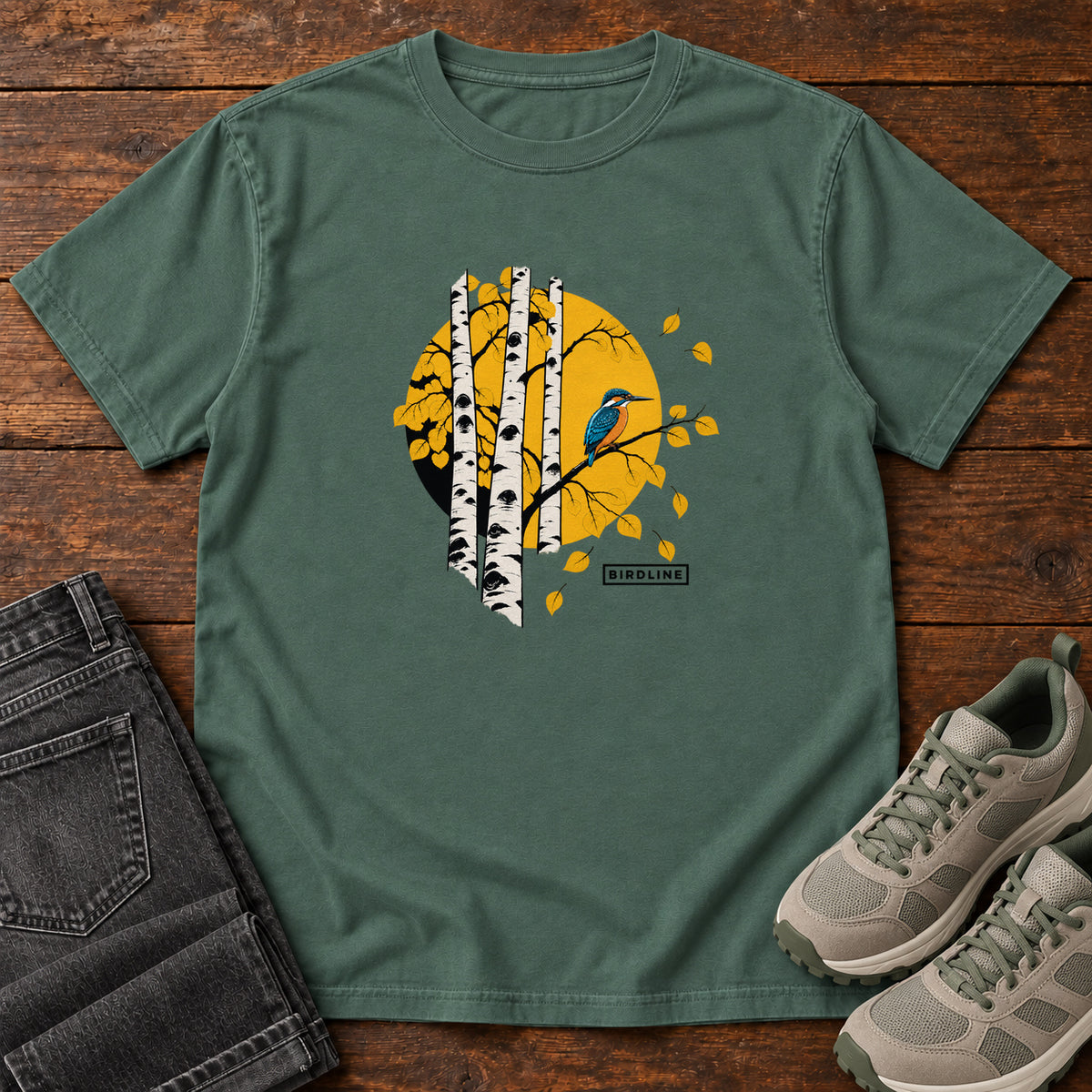 Blue Spruce Aspens Kingfisher T-Shirt with a blue bird perched among white tree trunks against a golden circle, front flat lay on wood.