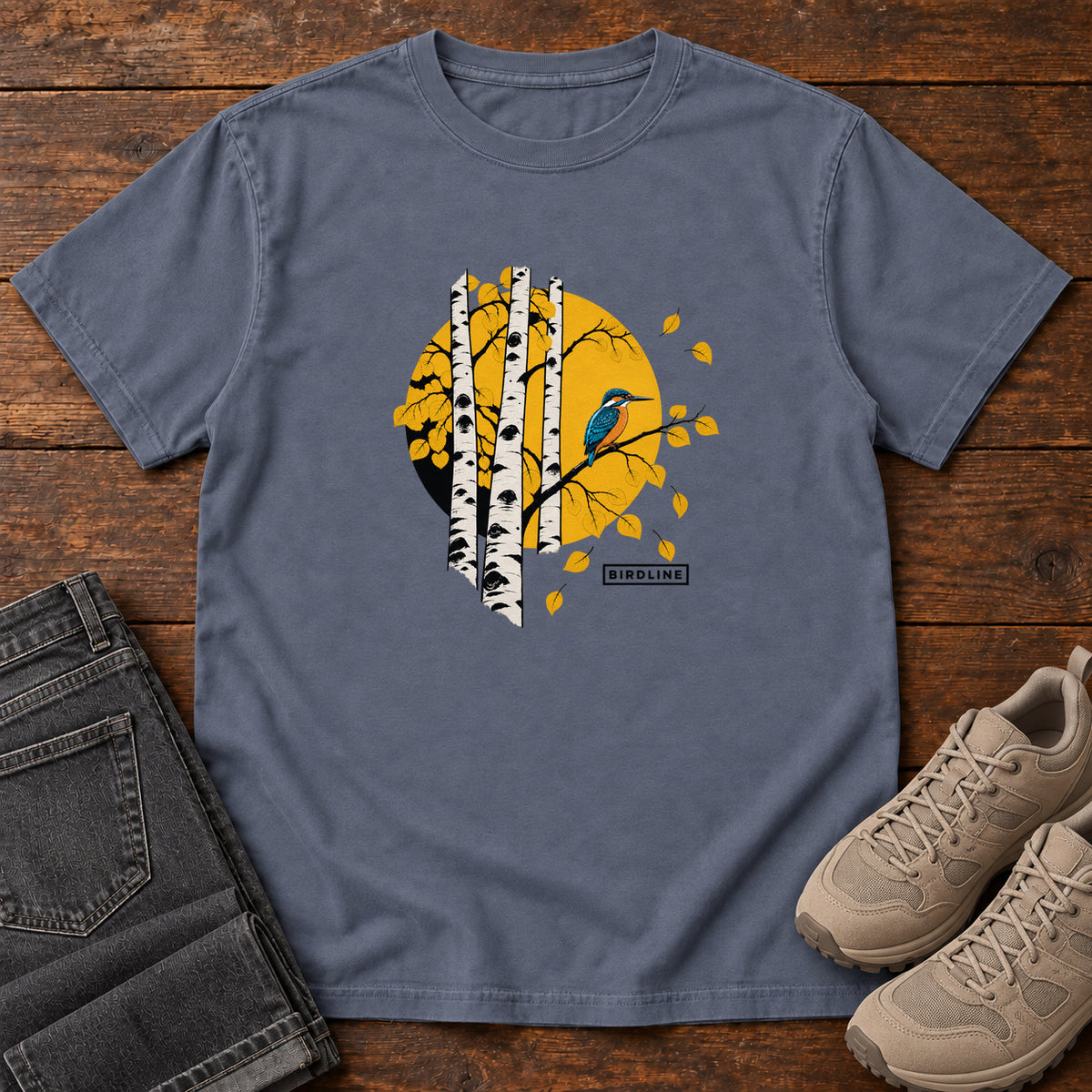 Denim Aspens Kingfisher T-Shirt with a blue bird perched among white tree trunks against a golden circle, front flat lay on wood.