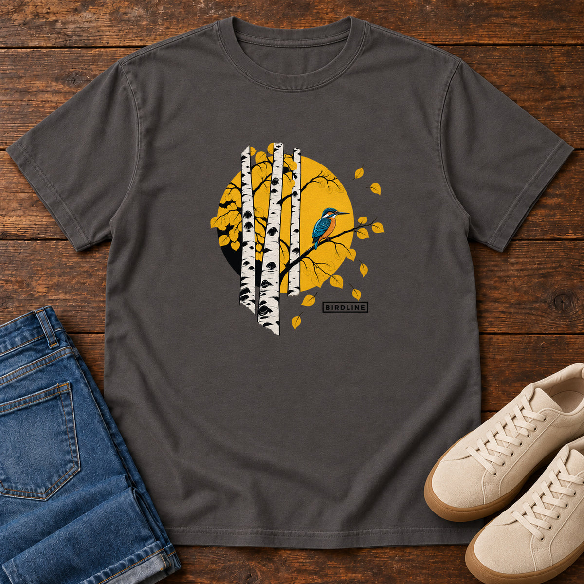 Graphite Aspens Kingfisher T-Shirt with a blue bird perched among white tree trunks against a golden circle, front flat lay on wood.