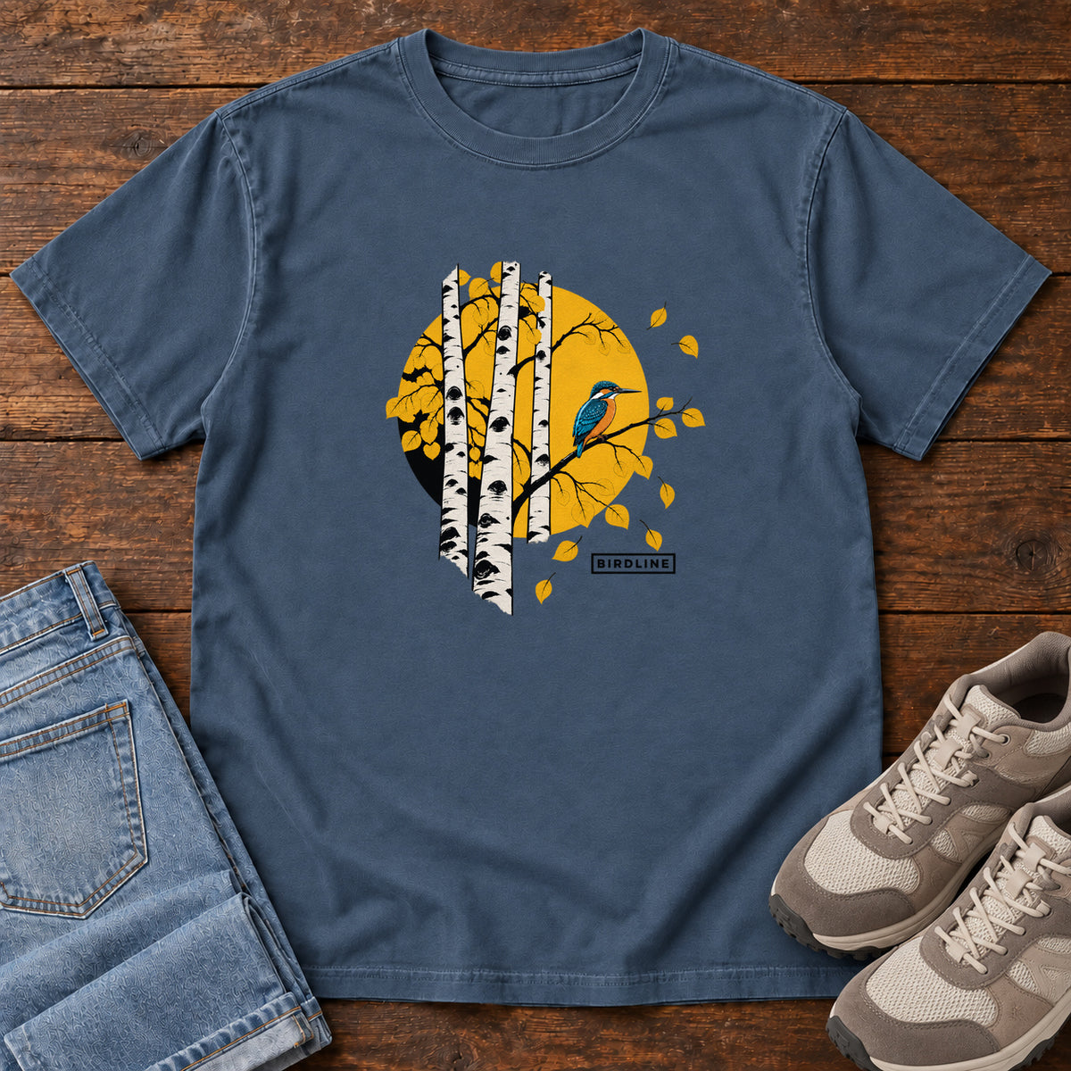 Midnight Aspens Kingfisher T-Shirt with a blue bird perched among white tree trunks against a golden circle, front flat lay on wood.