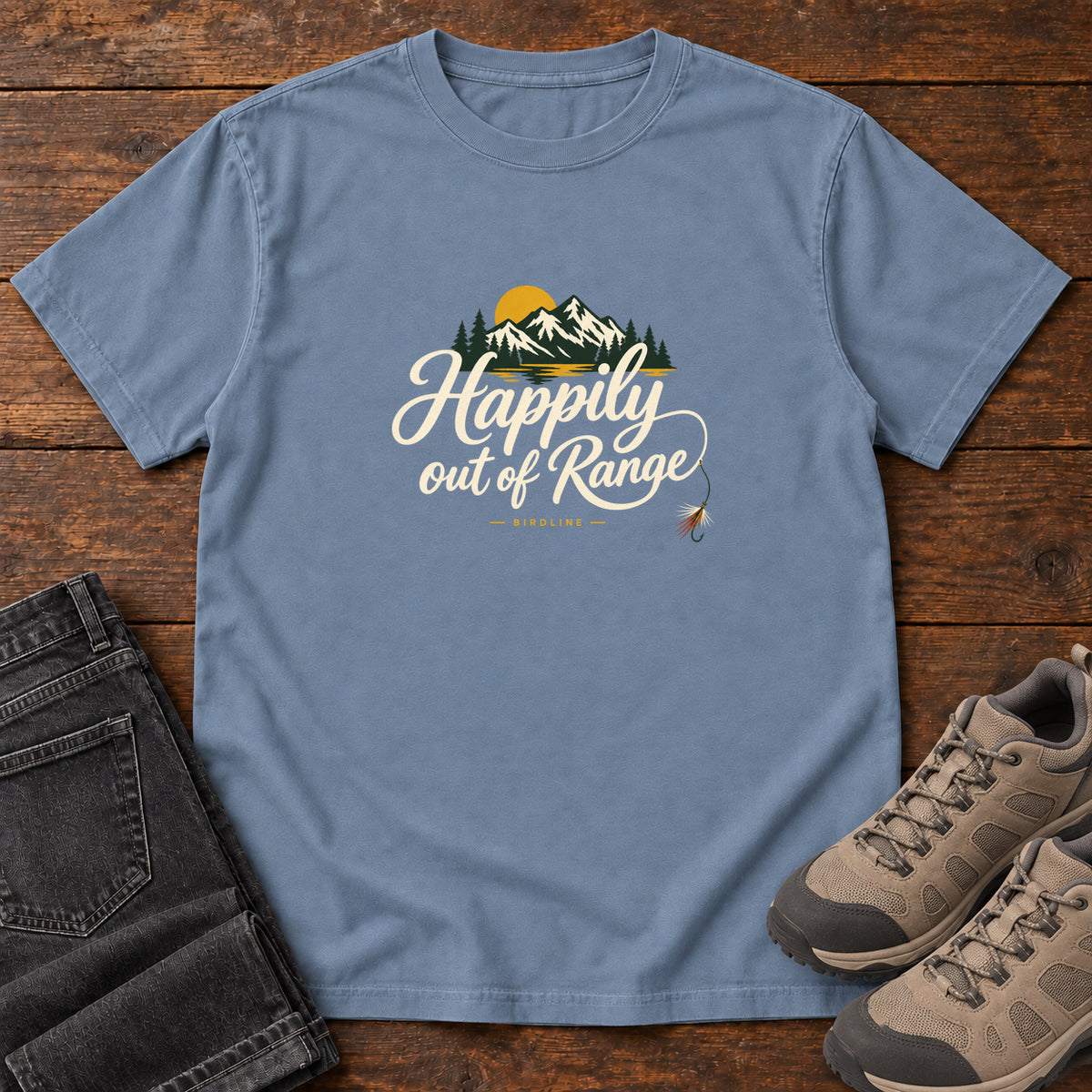 Blue Jean Happily out of Range T-Shirt with mountains, evergreens and a golden sun above cream script lettering; front flat lay on wood.