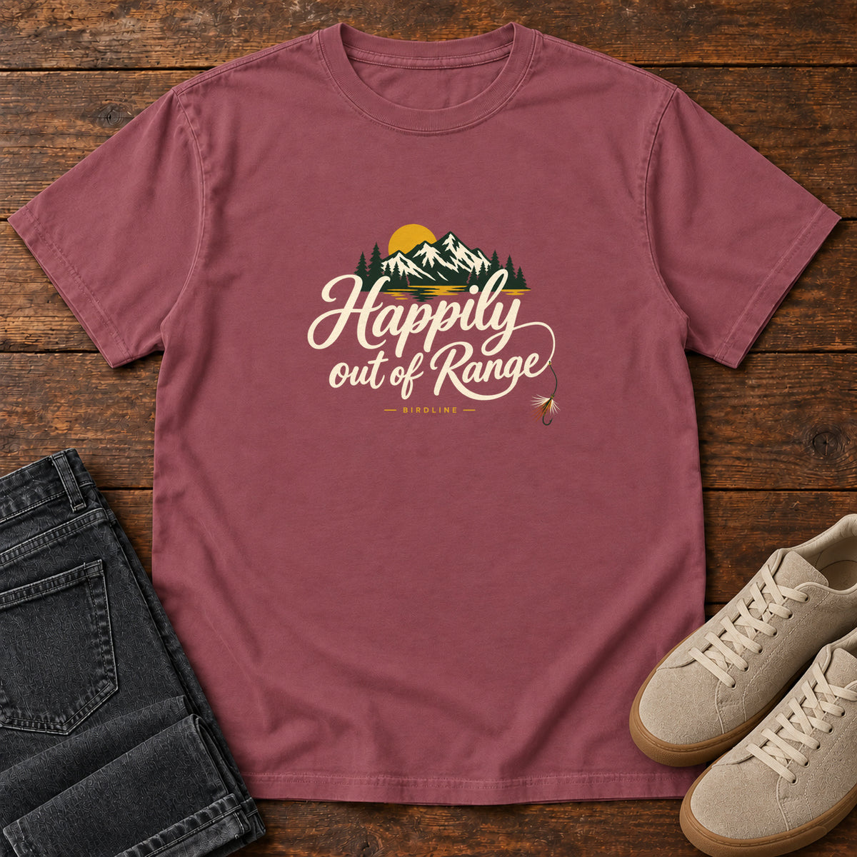 Brick Happily out of Range T-Shirt with mountains, evergreens and a golden sun above cream script lettering; front flat lay on wood.