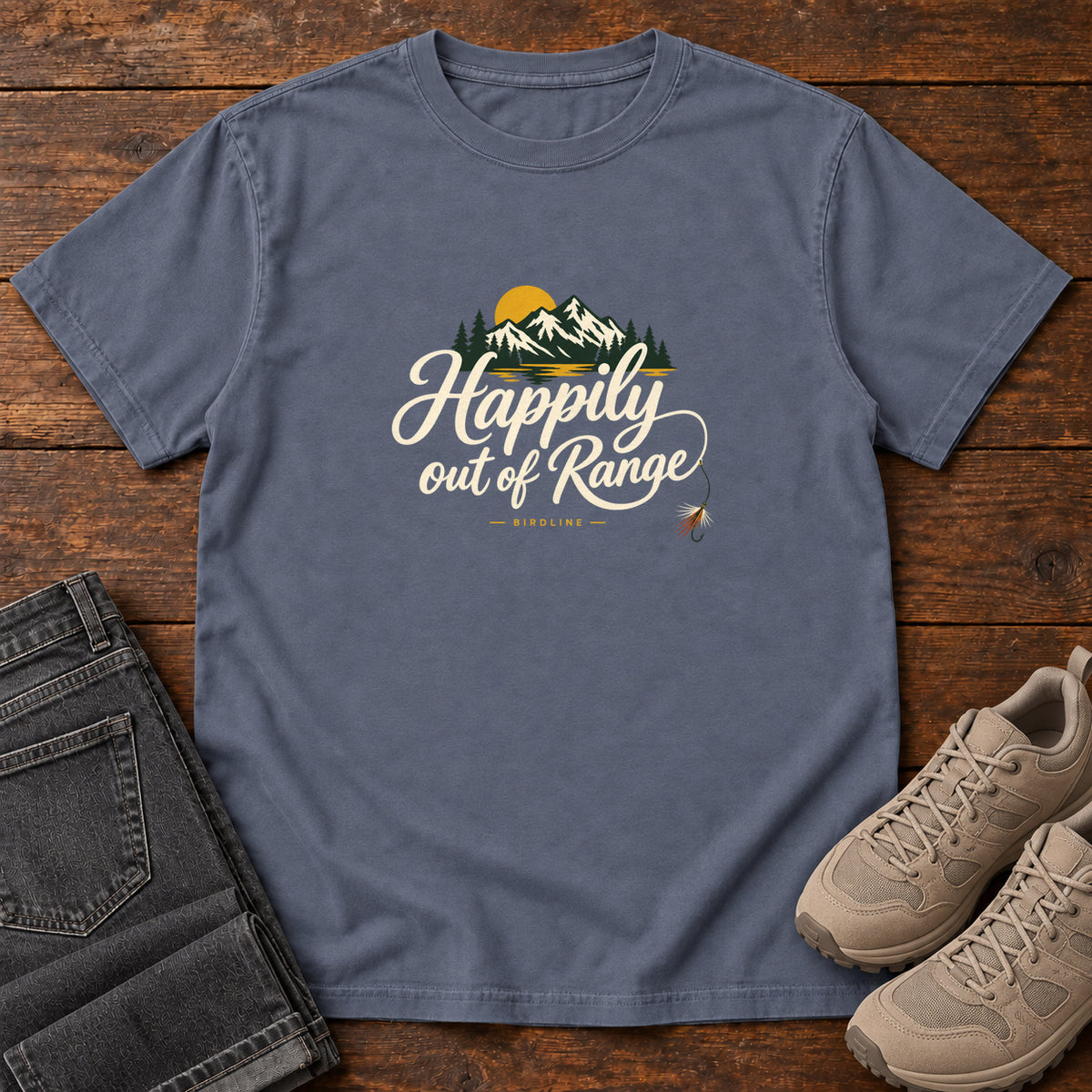 Denim Happily out of Range T-Shirt with mountains, evergreens and a golden sun above cream script lettering; front flat lay on wood.