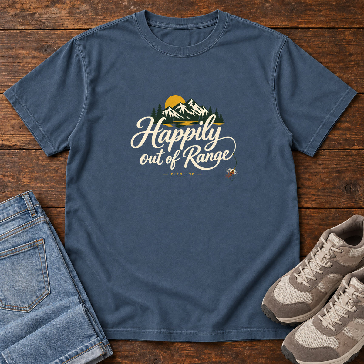 Midnight Happily out of Range T-Shirt with mountains, evergreens and a golden sun above cream script lettering; front flat lay on wood.