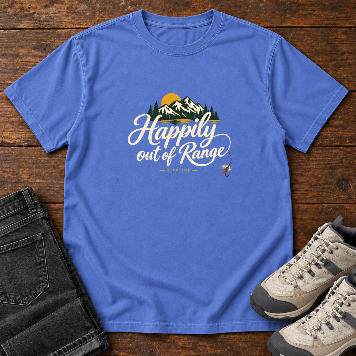 Mystic Blue Happily out of Range T-Shirt with mountains, evergreens and a golden sun above cream script lettering; front flat lay on wood.
