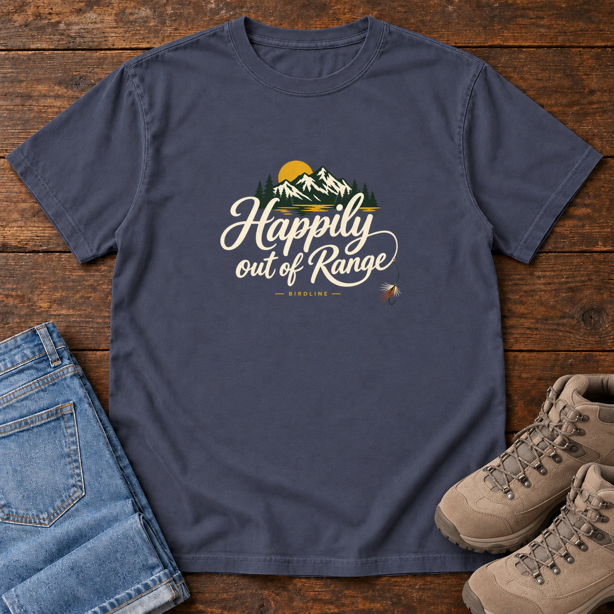 Navy Happily out of Range T-Shirt with mountains, evergreens and a golden sun above cream script lettering; front flat lay on wood.
