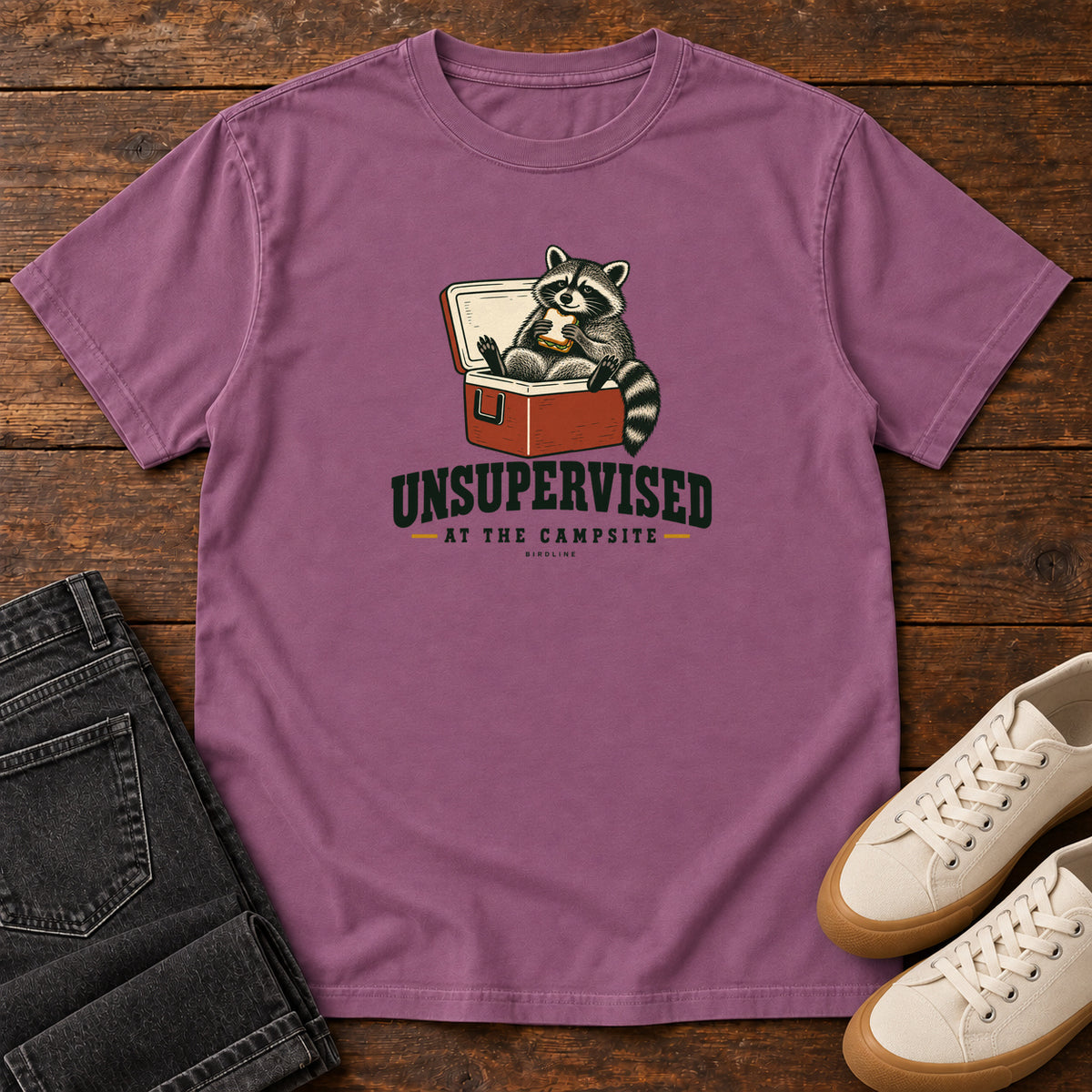 Berry Unsupervised at the Campsite T-Shirt with a raccoon eating a sandwich while sitting in an open red cooler; front flat lay on wood.