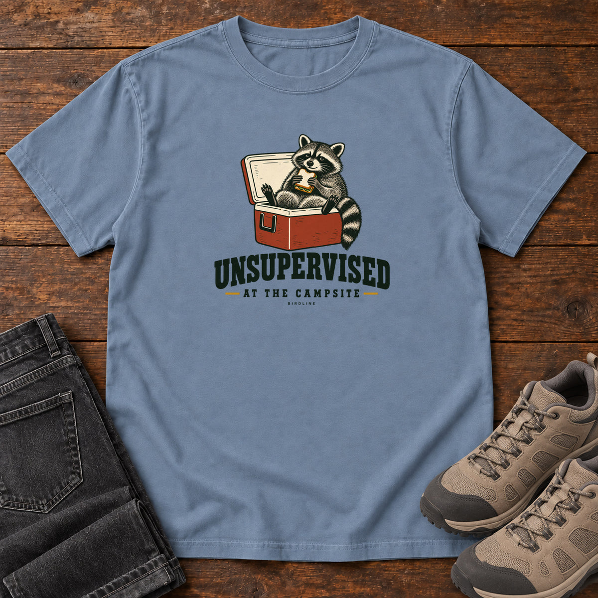Blue Jean Unsupervised at the Campsite T-Shirt with a raccoon eating a sandwich while sitting in an open red cooler; front flat lay on wood.