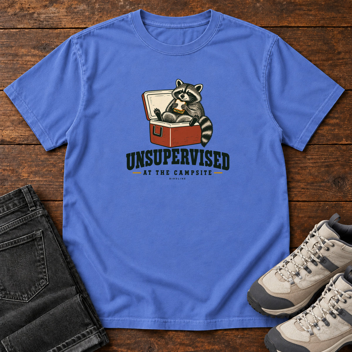 Mystic Blue Unsupervised at the Campsite T-Shirt with a raccoon eating a sandwich while sitting in an open red cooler; front flat lay on wood.