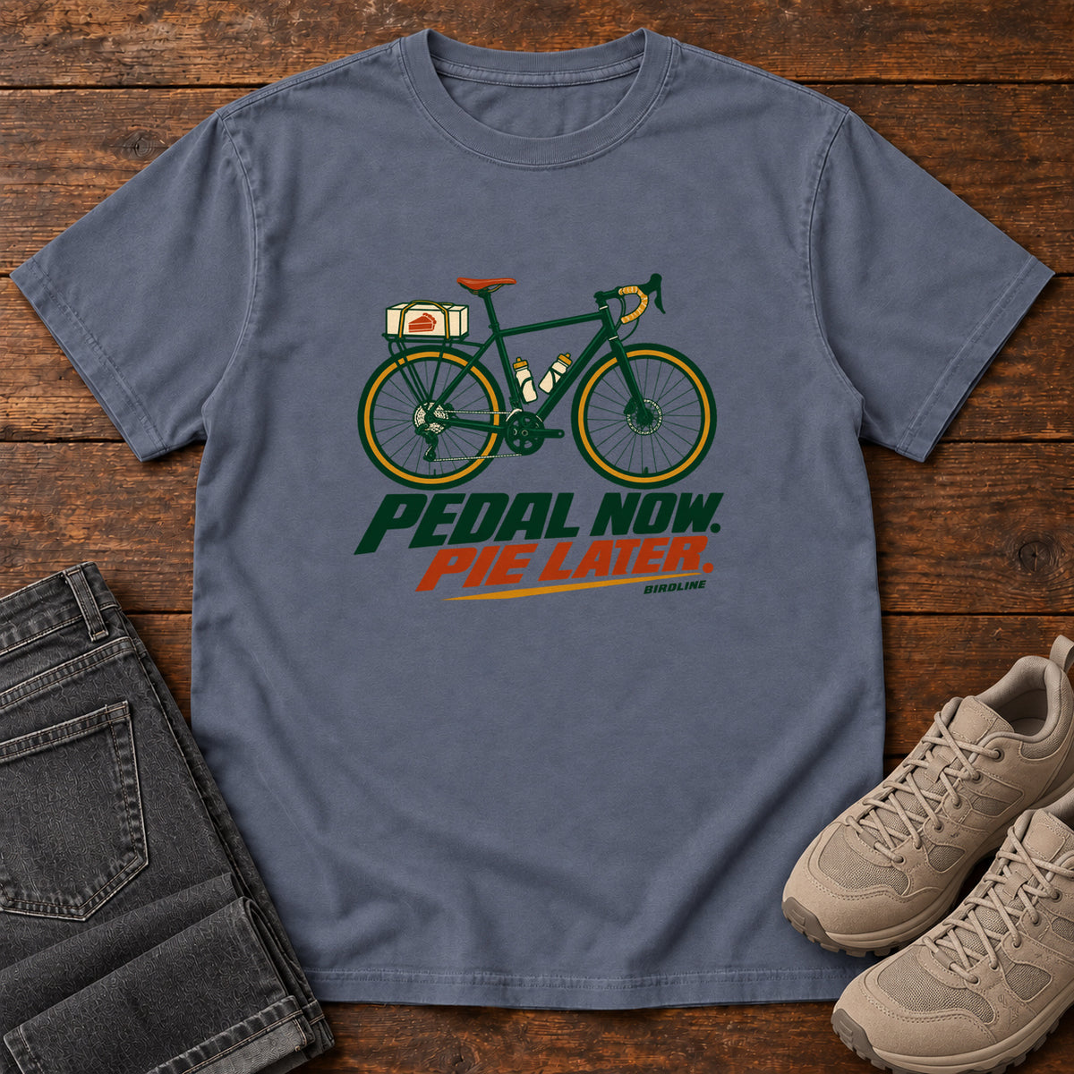 Denim Pedal Now Pie Later T-Shirt with a green touring bicycle above green and orange lettering, front flat lay on wood.