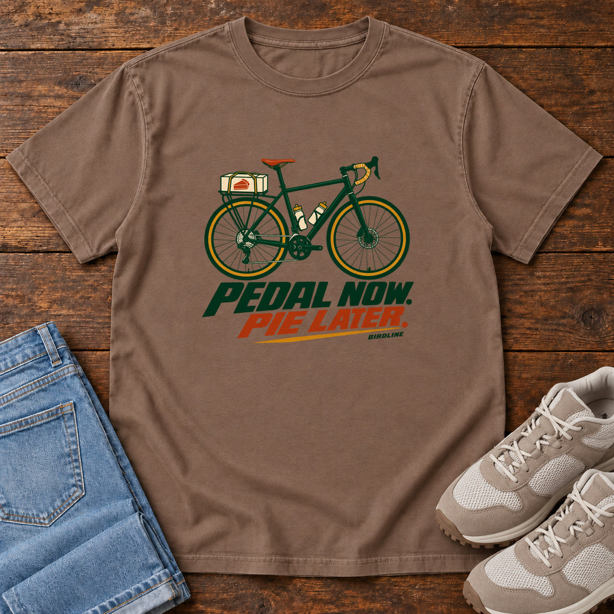 Espresso Pedal Now Pie Later T-Shirt with a green touring bicycle above green and orange lettering, front flat lay on wood.