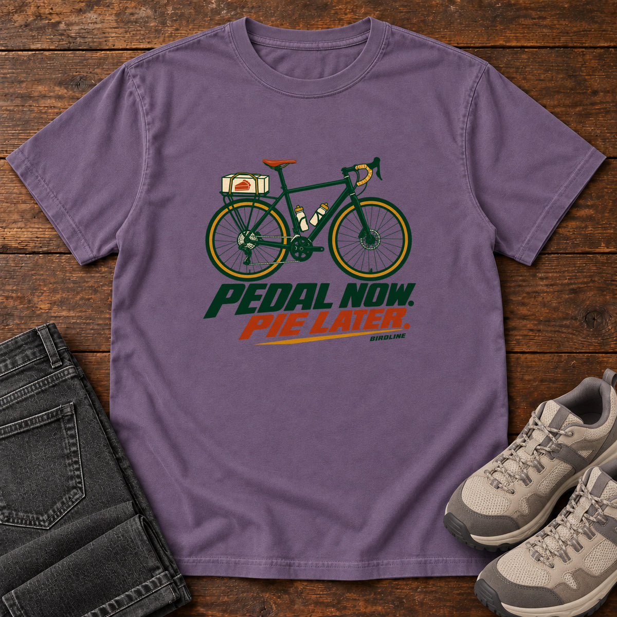 Grape Pedal Now Pie Later T-Shirt with a green touring bicycle above green and orange lettering, front flat lay on wood.