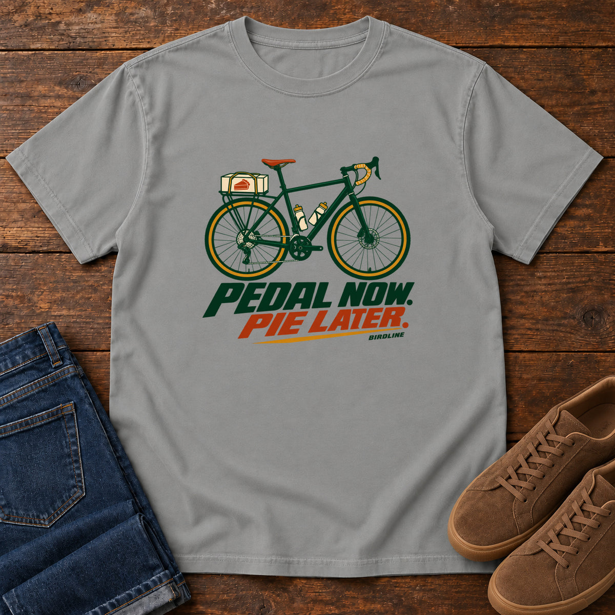 Grey Pedal Now Pie Later T-Shirt with a green touring bicycle above green and orange lettering, front flat lay on wood.