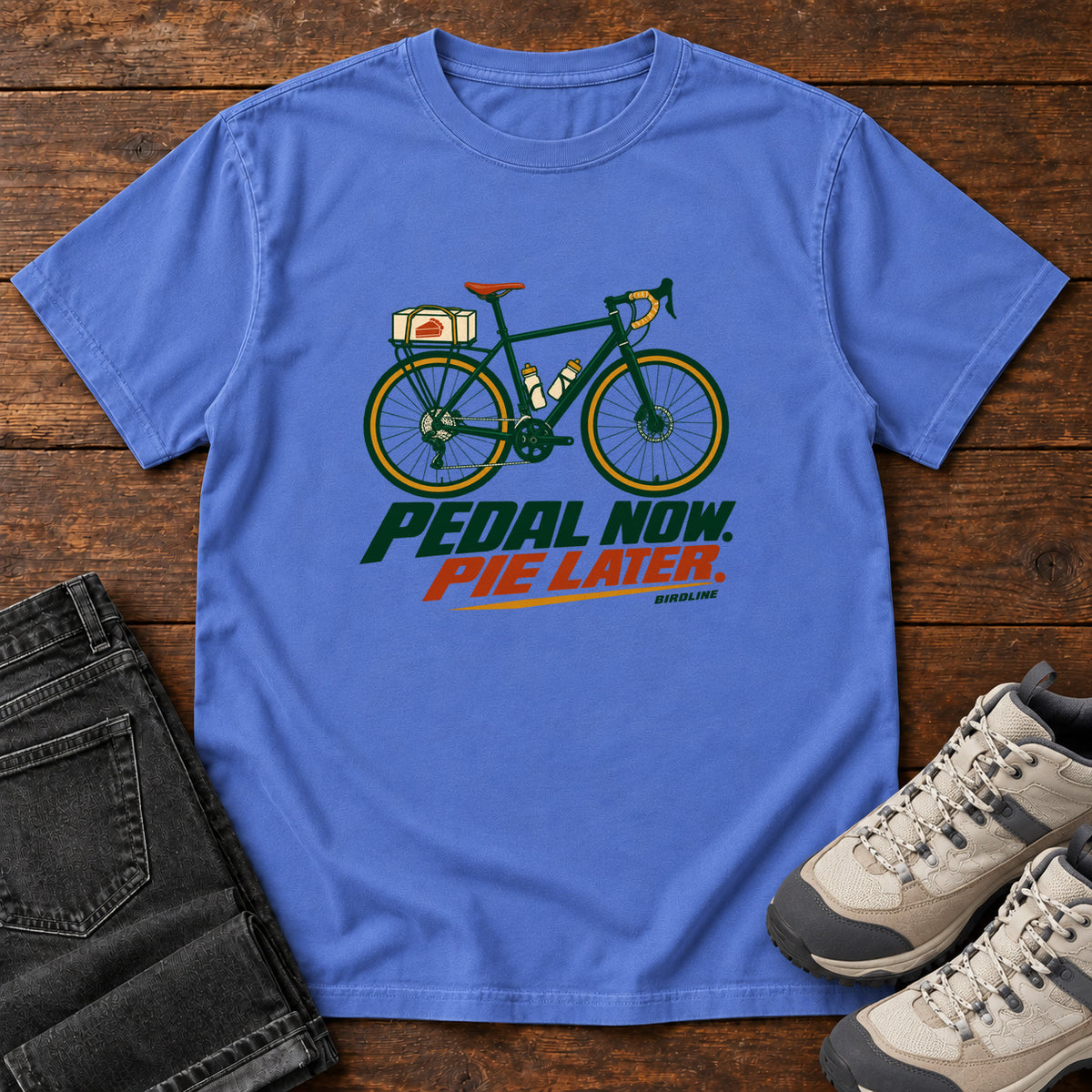 Mystic Blue Pedal Now Pie Later T-Shirt with a green touring bicycle above green and orange lettering, front flat lay on wood.