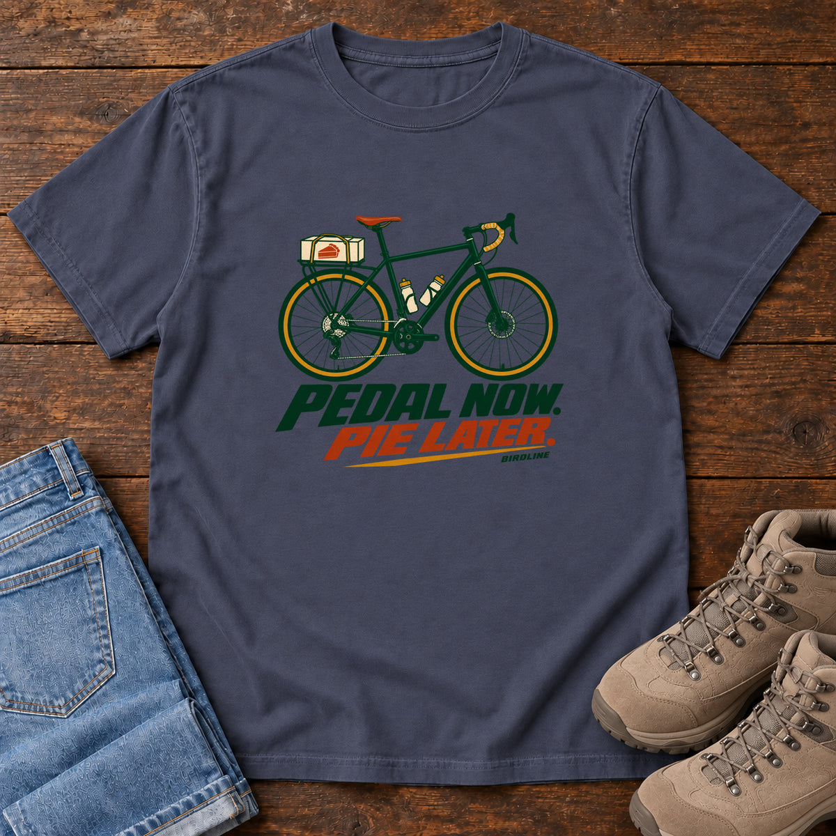 Navy Pedal Now Pie Later T-Shirt with a green touring bicycle above green and orange lettering, front flat lay on wood.
