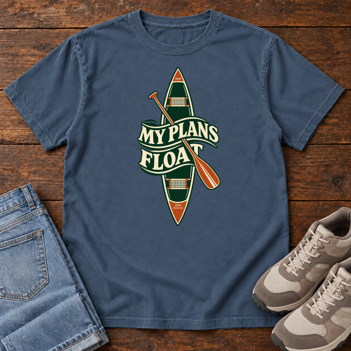 My Plans Float T-Shirt in Midnight, front laid flat on wood, featuring a vertical green canoe and orange paddle behind cream slogan lettering.