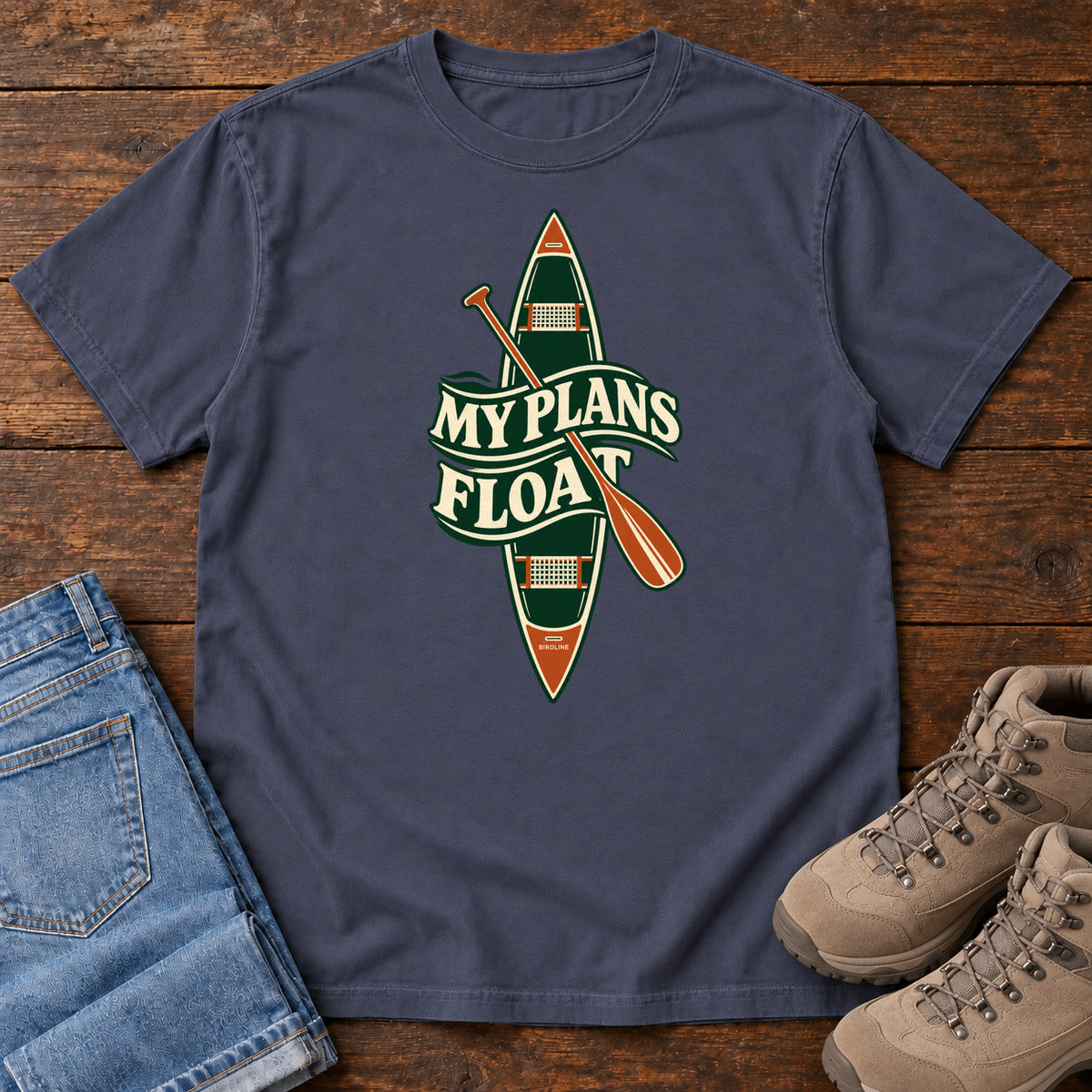 My Plans Float T-Shirt in Navy, front laid flat on wood, featuring a vertical green canoe and orange paddle behind cream slogan lettering.