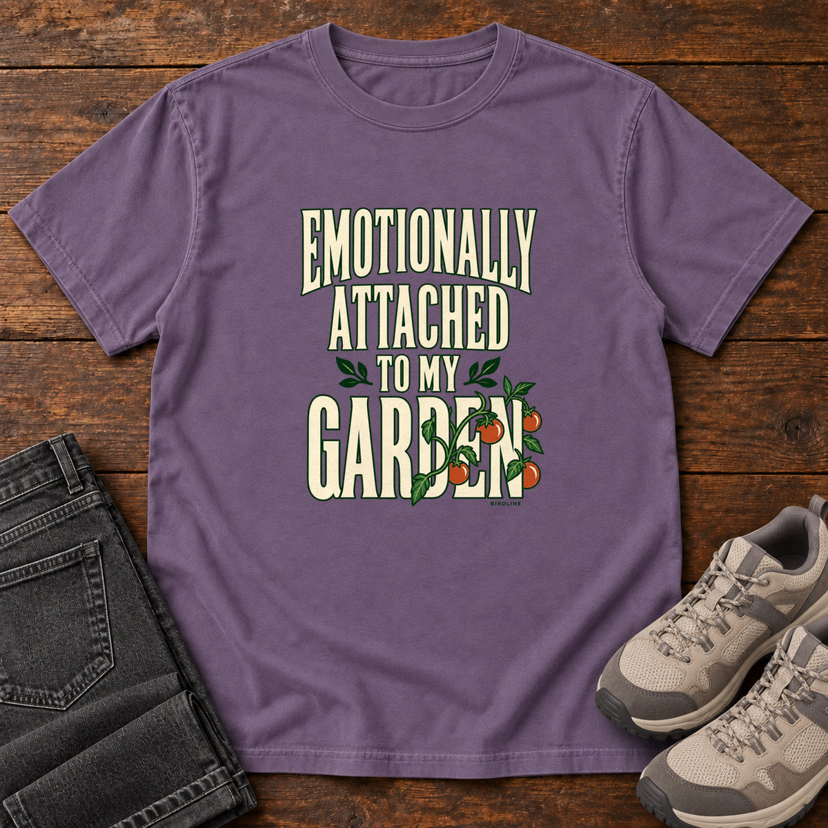 Grape Emotionally Attached to My Garden T-Shirt with cream lettering intertwined with a leafy vine and orange fruit, front flat lay on wood.