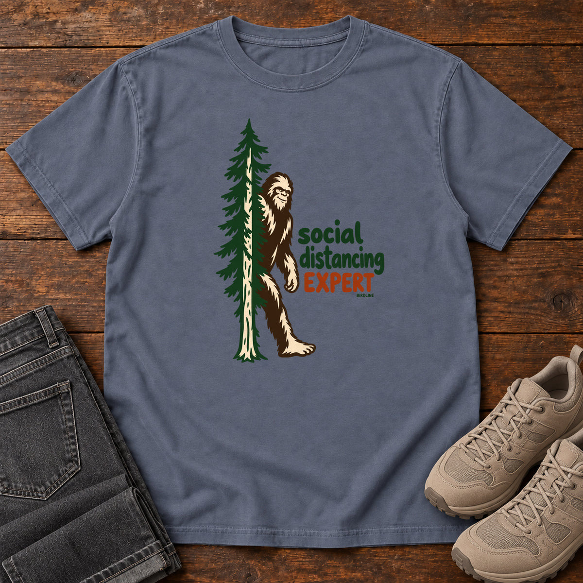 Denim Social Distancing Expert T-Shirt with Bigfoot beside an evergreen and Social Distancing Expert lettering; front flat lay on wood.