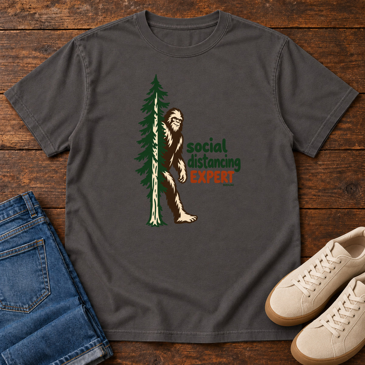 Graphite Social Distancing Expert T-Shirt with Bigfoot beside an evergreen and Social Distancing Expert lettering; front flat lay on wood.