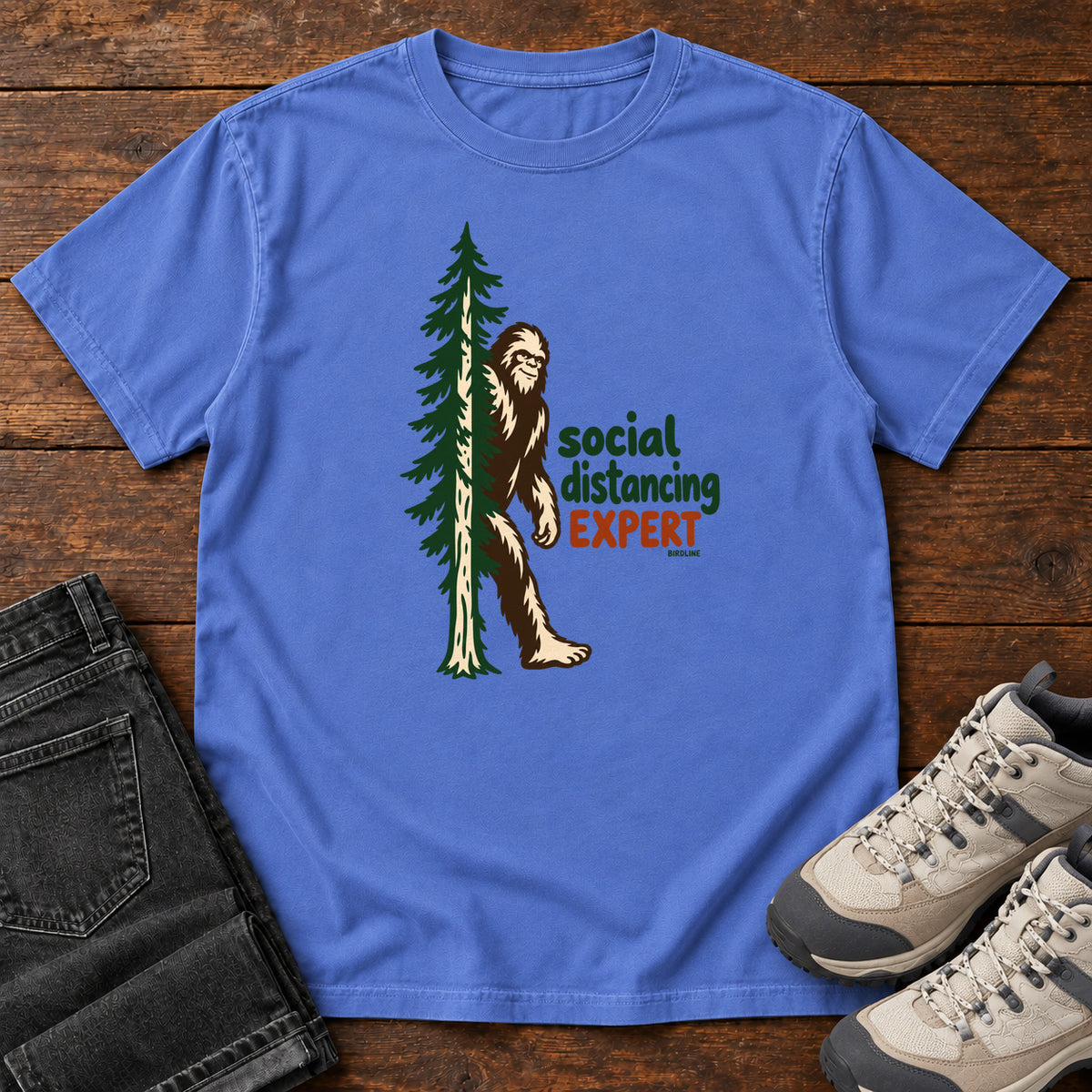 Mystic Blue Social Distancing Expert T-Shirt with Bigfoot beside an evergreen and Social Distancing Expert lettering; front flat lay on wood.