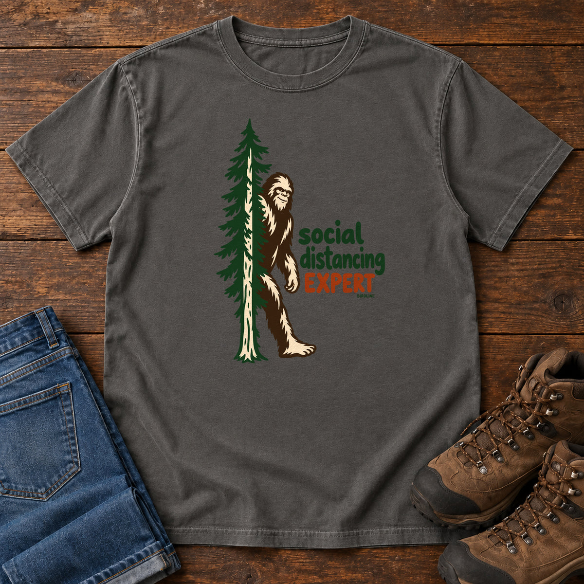 Pepper Social Distancing Expert T-Shirt with Bigfoot beside an evergreen and Social Distancing Expert lettering; front flat lay on wood.