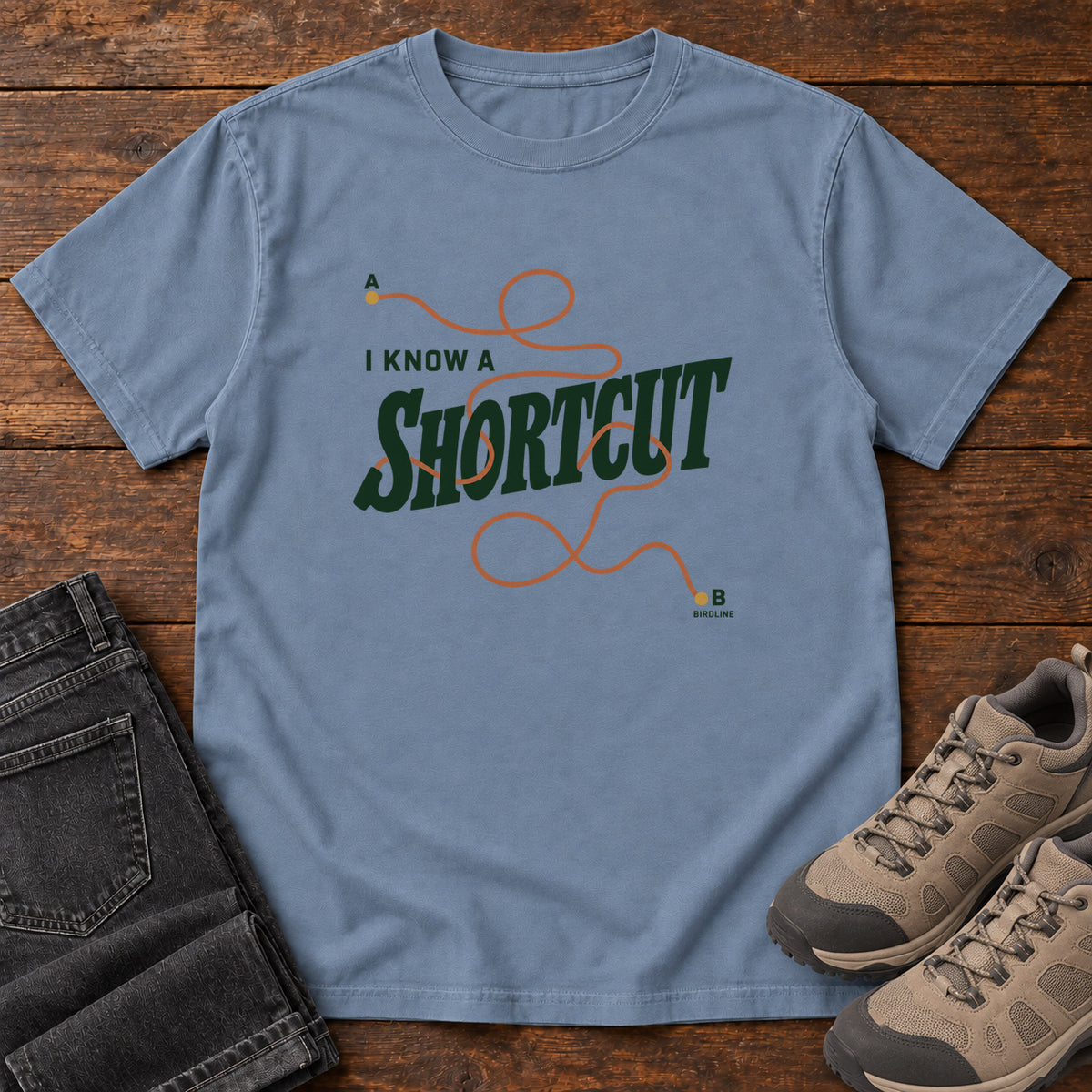 I Know a Shortcut T-Shirt in Blue Jean, front laid flat on wood, featuring dark green slogan lettering crossed by a looping orange route line.