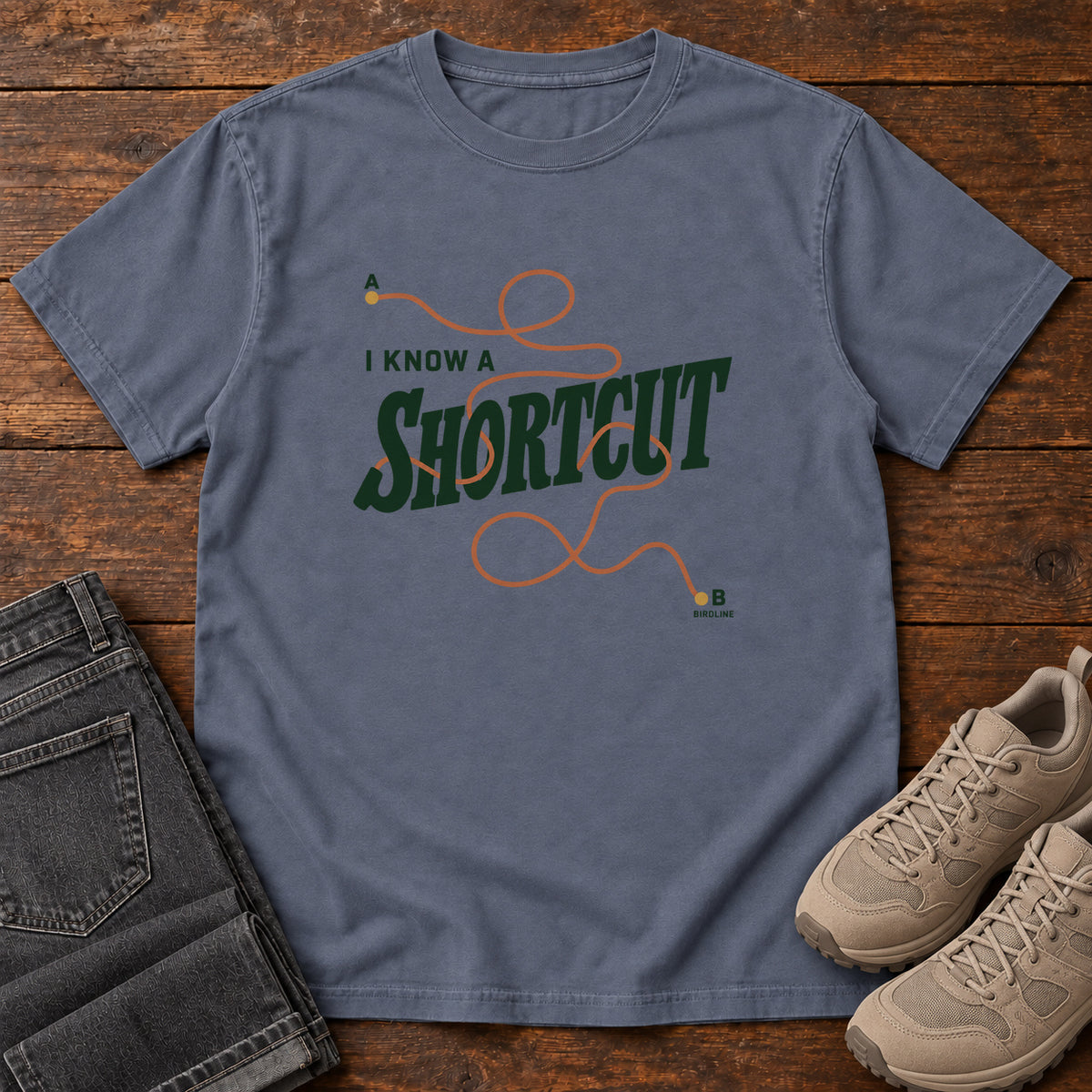 I Know a Shortcut T-Shirt in Denim, front laid flat on wood, featuring dark green slogan lettering crossed by a looping orange route line.