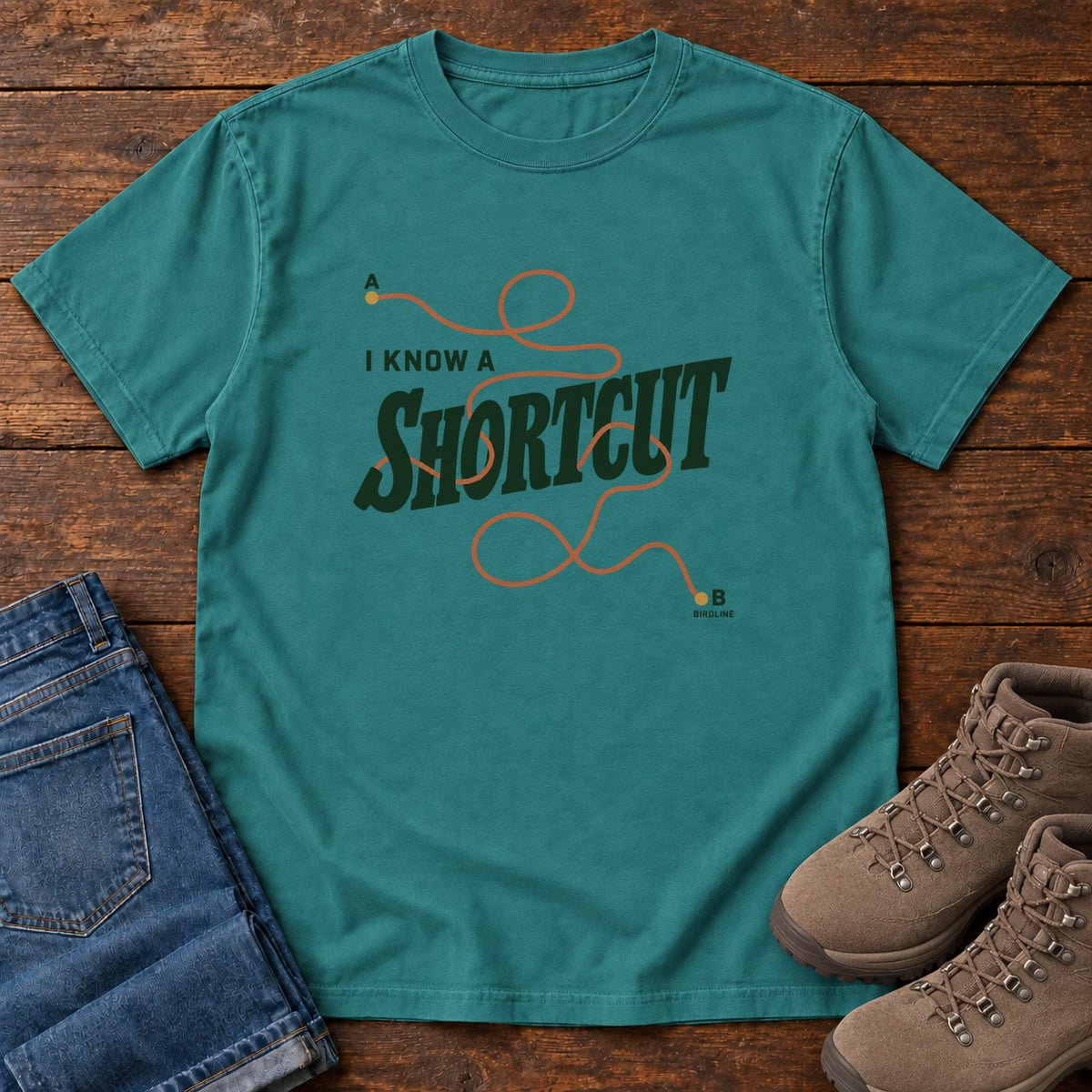 I Know a Shortcut T-Shirt in Emerald, front laid flat on wood, featuring dark green slogan lettering crossed by a looping orange route line.