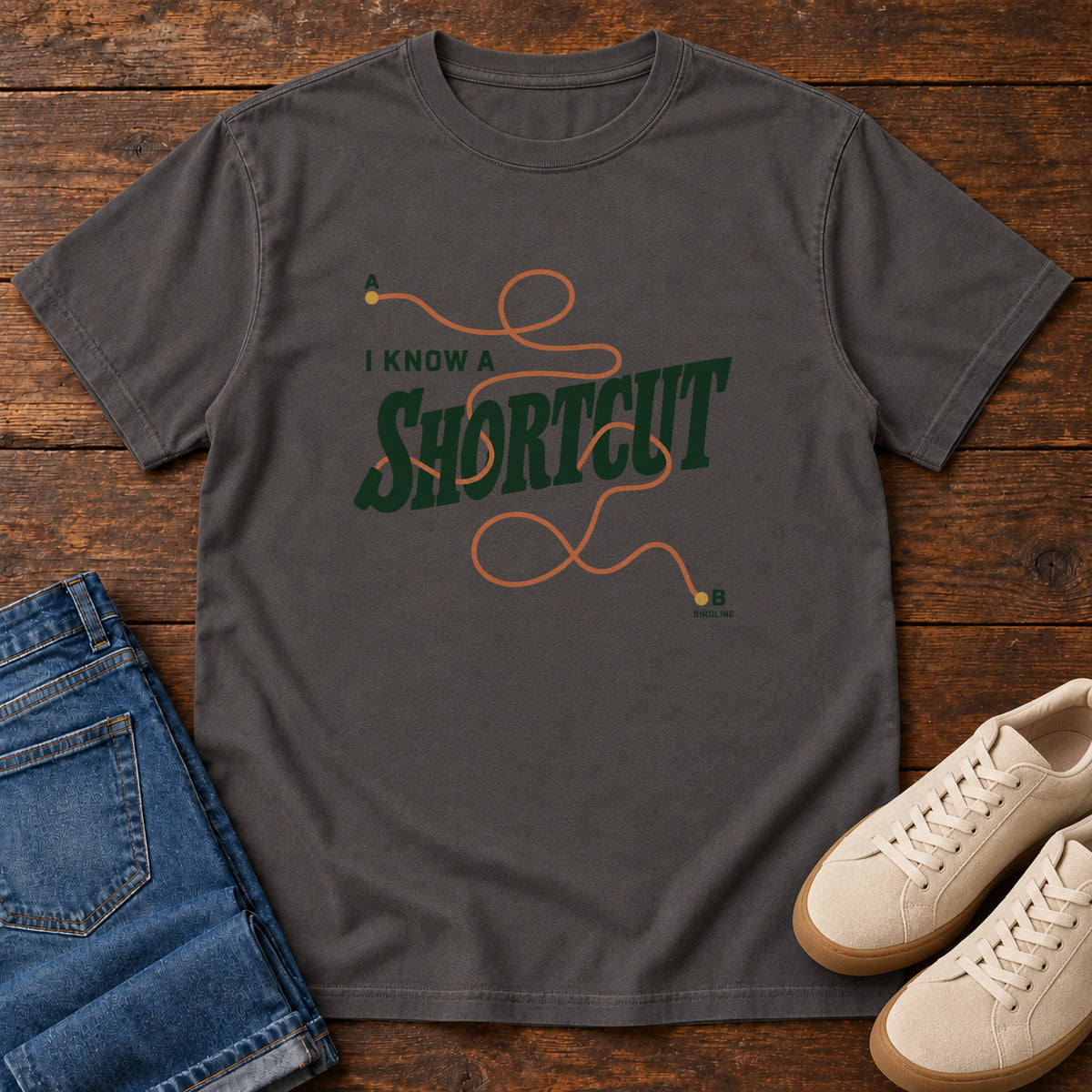 I Know a Shortcut T-Shirt in Graphite, front laid flat on wood, featuring dark green slogan lettering crossed by a looping orange route line.
