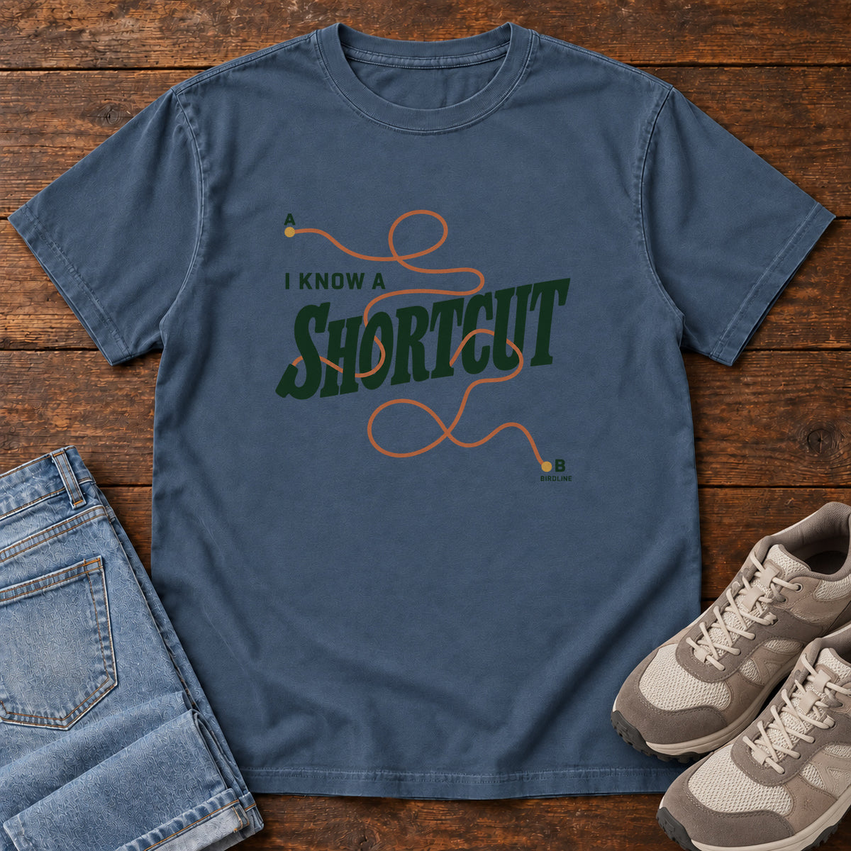 I Know a Shortcut T-Shirt in Midnight, front laid flat on wood, featuring dark green slogan lettering crossed by a looping orange route line.