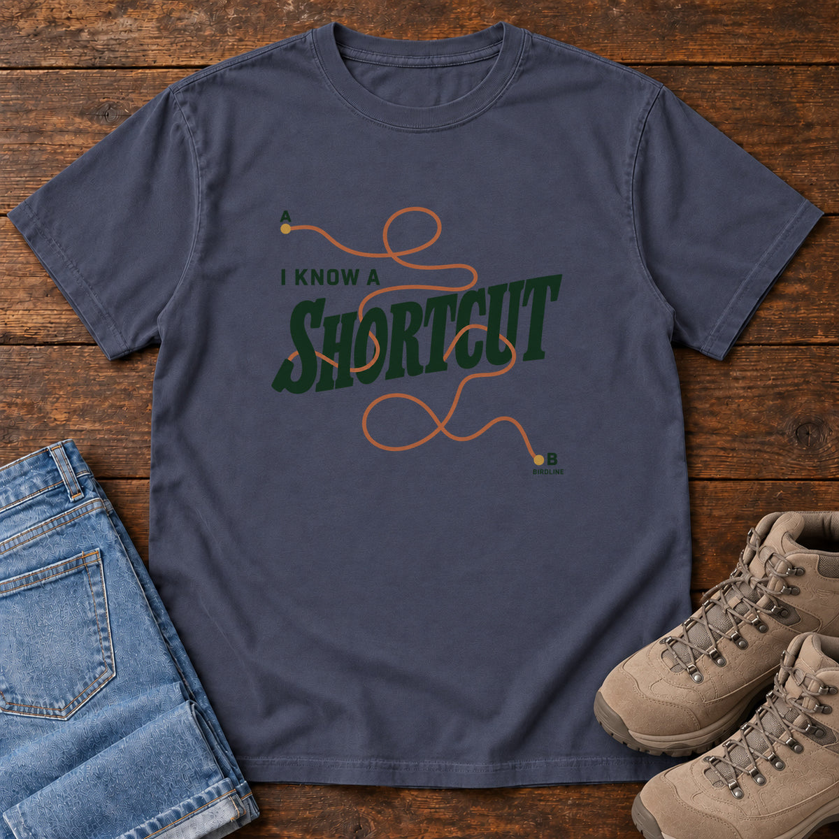 I Know a Shortcut T-Shirt in Navy, front laid flat on wood, featuring dark green slogan lettering crossed by a looping orange route line.