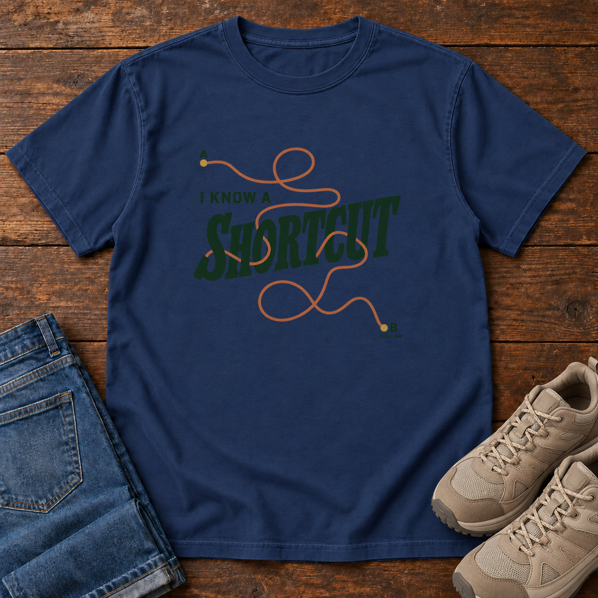 I Know a Shortcut T-Shirt in True Navy, front laid flat on wood, featuring dark green slogan lettering crossed by a looping orange route line.
