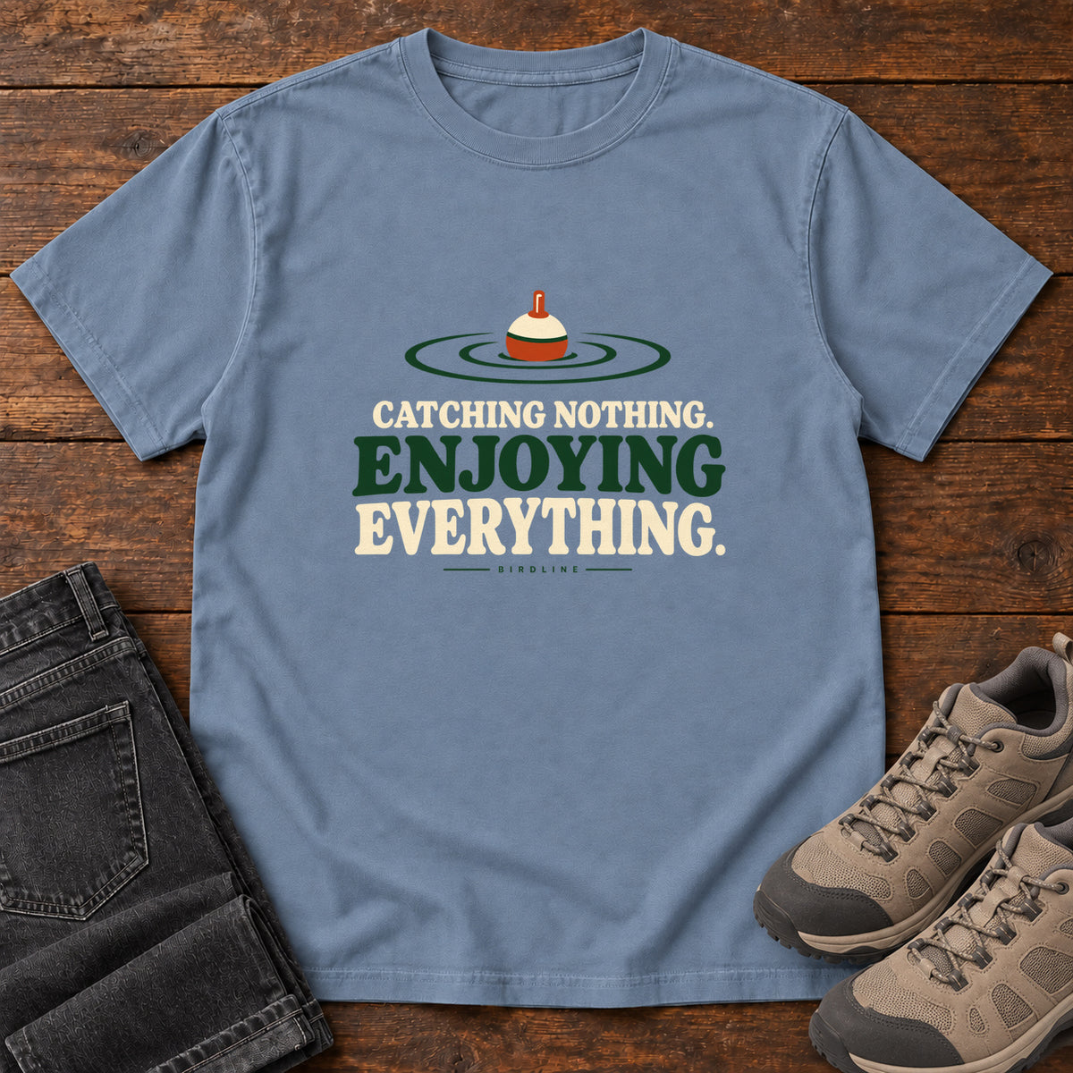 Blue Jean Catching Nothing Enjoying Everything T-Shirt with a red-and-cream fishing bobber in green ripples above bold lettering; front flat lay on wood.