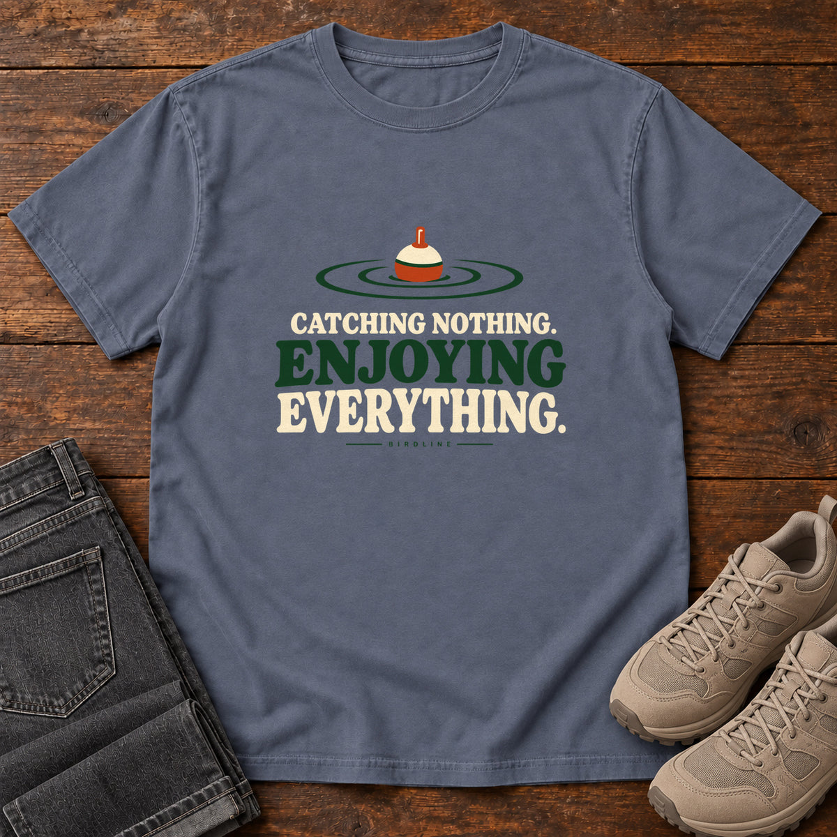 Denim Catching Nothing Enjoying Everything T-Shirt with a red-and-cream fishing bobber in green ripples above bold lettering; front flat lay on wood.
