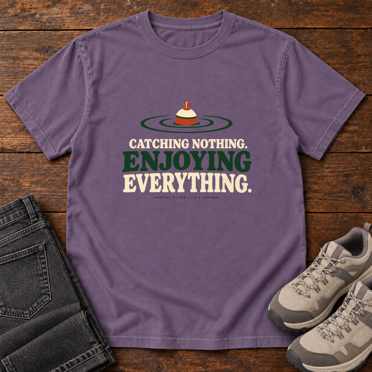 Grape Catching Nothing Enjoying Everything T-Shirt with a red-and-cream fishing bobber in green ripples above bold lettering; front flat lay on wood.