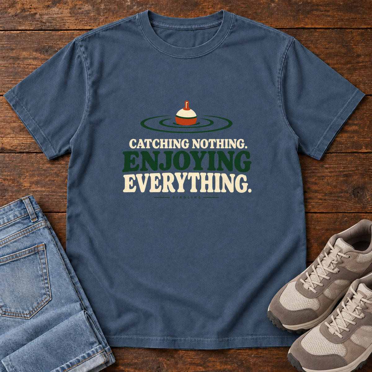 Midnight Catching Nothing Enjoying Everything T-Shirt with a red-and-cream fishing bobber in green ripples above bold lettering; front flat lay on wood.