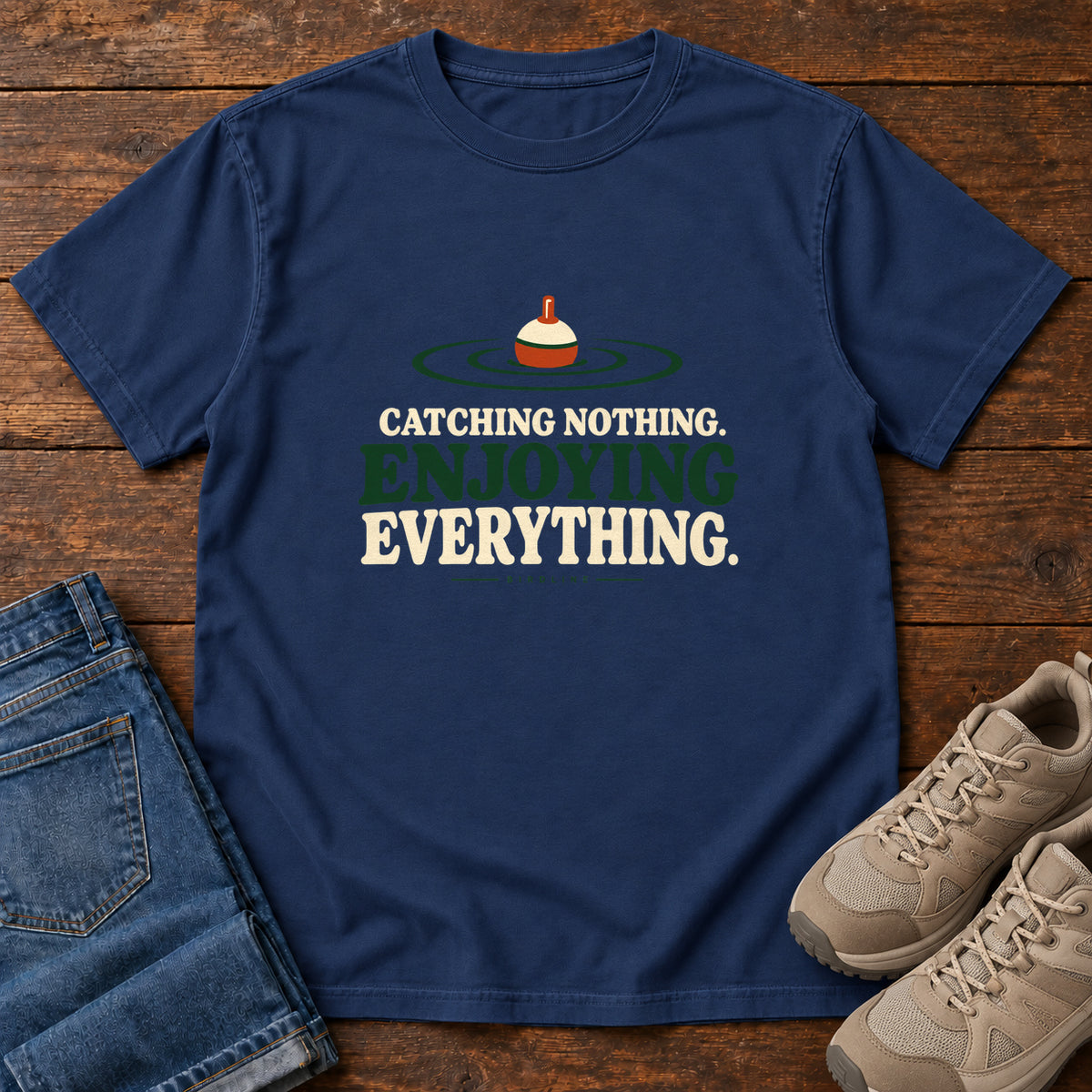 True Navy Catching Nothing Enjoying Everything T-Shirt with a red-and-cream fishing bobber in green ripples above bold lettering; front flat lay on wood.