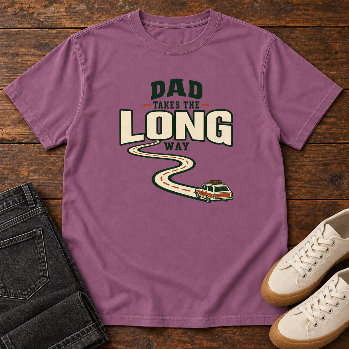 Berry Dad Takes the Long Way T-Shirt with a winding road leading to a station wagon beneath bold cream and green lettering, front flat lay on wood.