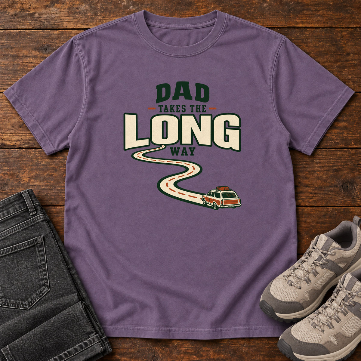 Grape Dad Takes the Long Way T-Shirt with a winding road leading to a station wagon beneath bold cream and green lettering, front flat lay on wood.