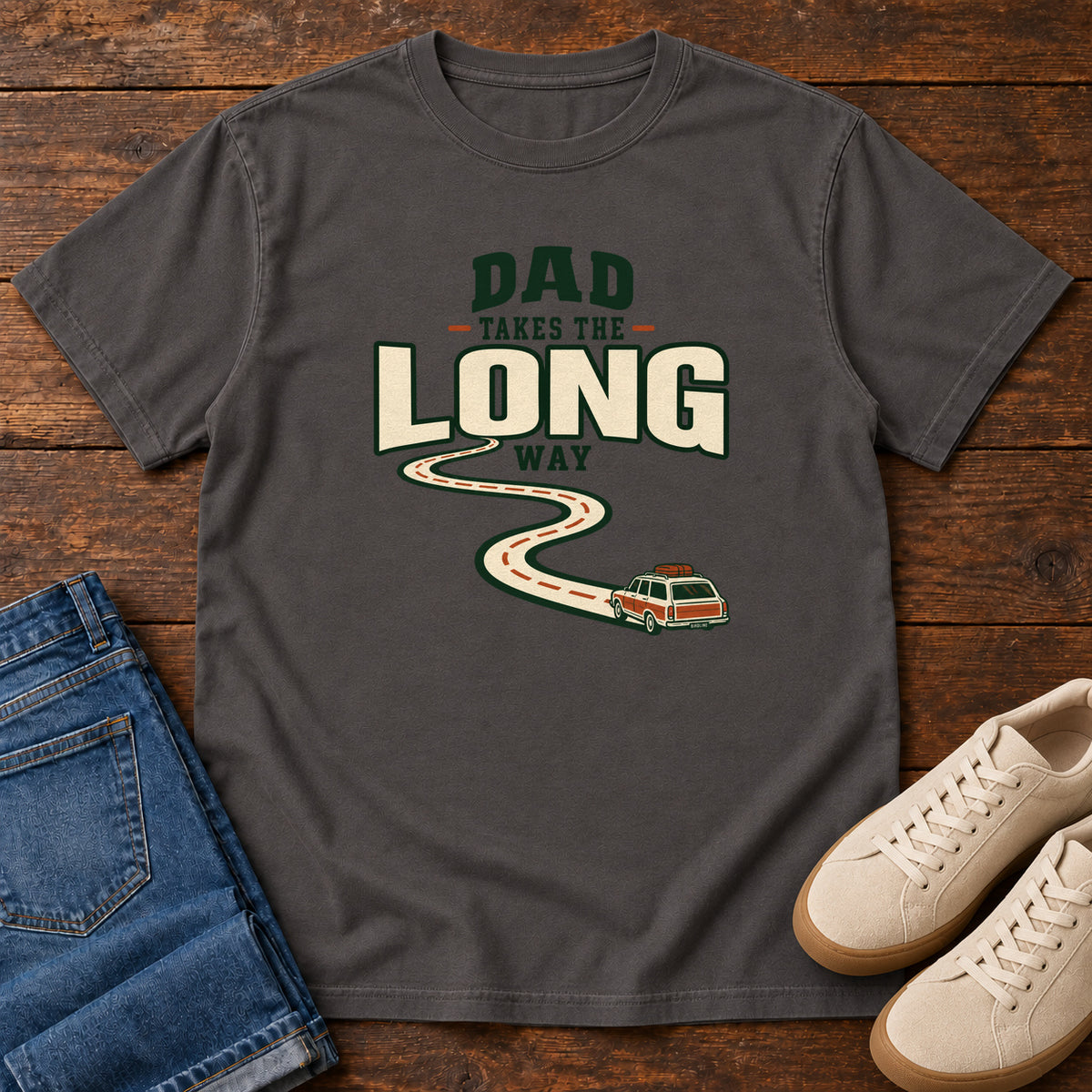 Graphite Dad Takes the Long Way T-Shirt with a winding road leading to a station wagon beneath bold cream and green lettering, front flat lay on wood.