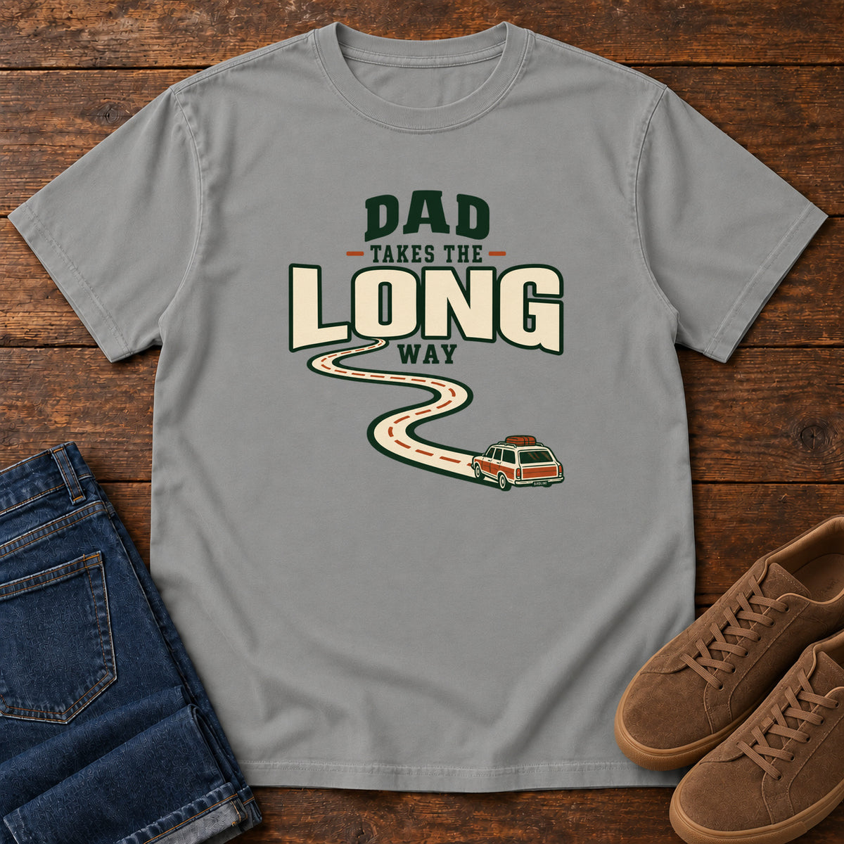 Grey Dad Takes the Long Way T-Shirt with a winding road leading to a station wagon beneath bold cream and green lettering, front flat lay on wood.