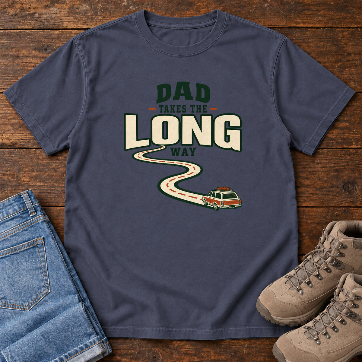 Navy Dad Takes the Long Way T-Shirt with a winding road leading to a station wagon beneath bold cream and green lettering, front flat lay on wood.