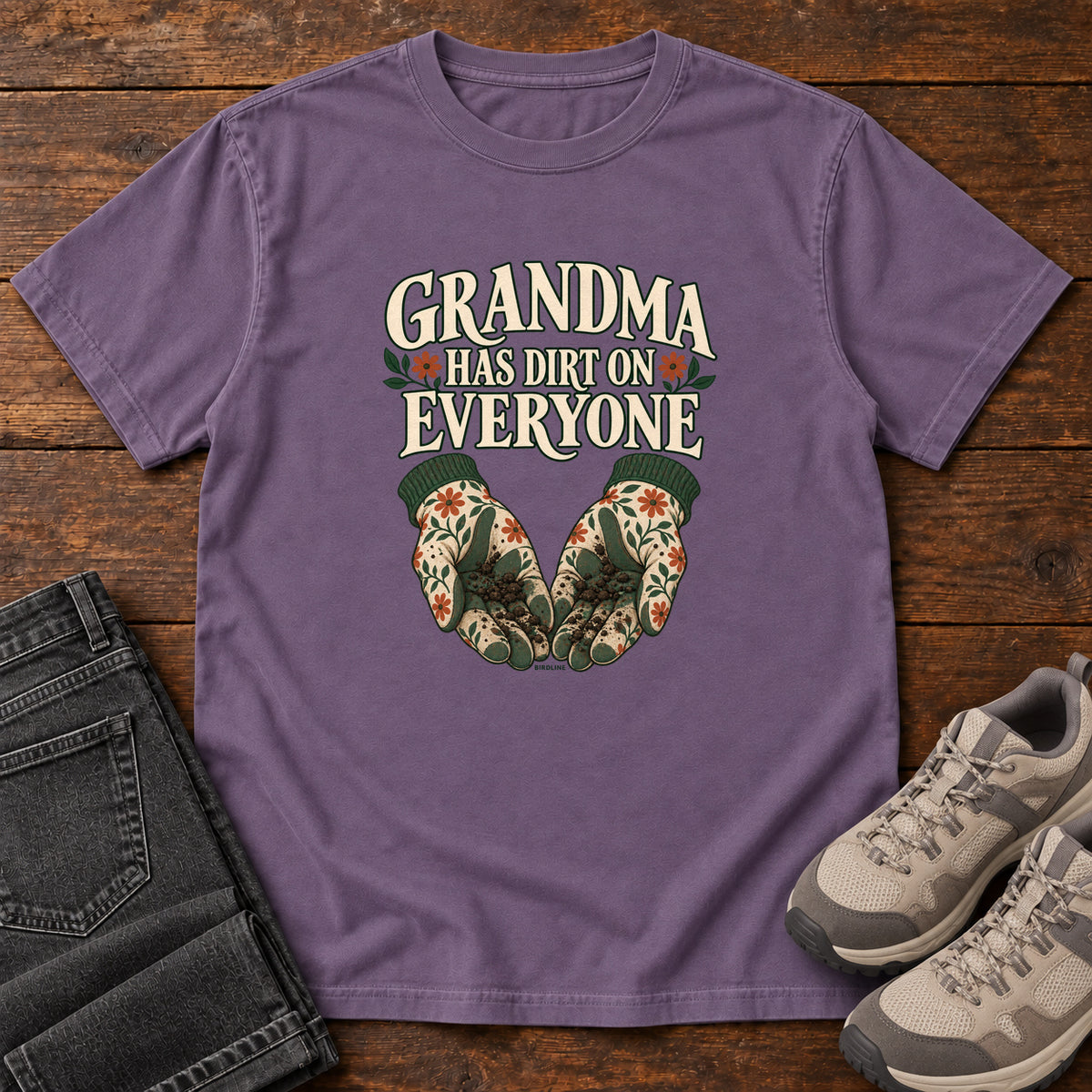 Grandma Has Dirt on Everyone T-Shirt in Grape, front laid flat on wood, featuring floral gardening gloves cupping soil beneath large cream slogan lettering.
