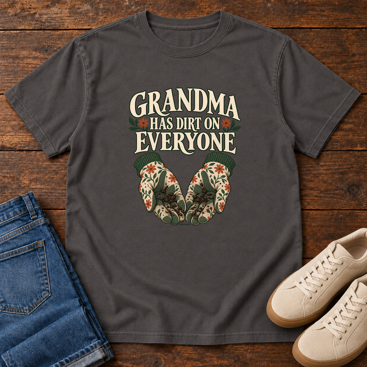 Grandma Has Dirt on Everyone T-Shirt in Graphite, front laid flat on wood, featuring floral gardening gloves cupping soil beneath large cream slogan lettering.