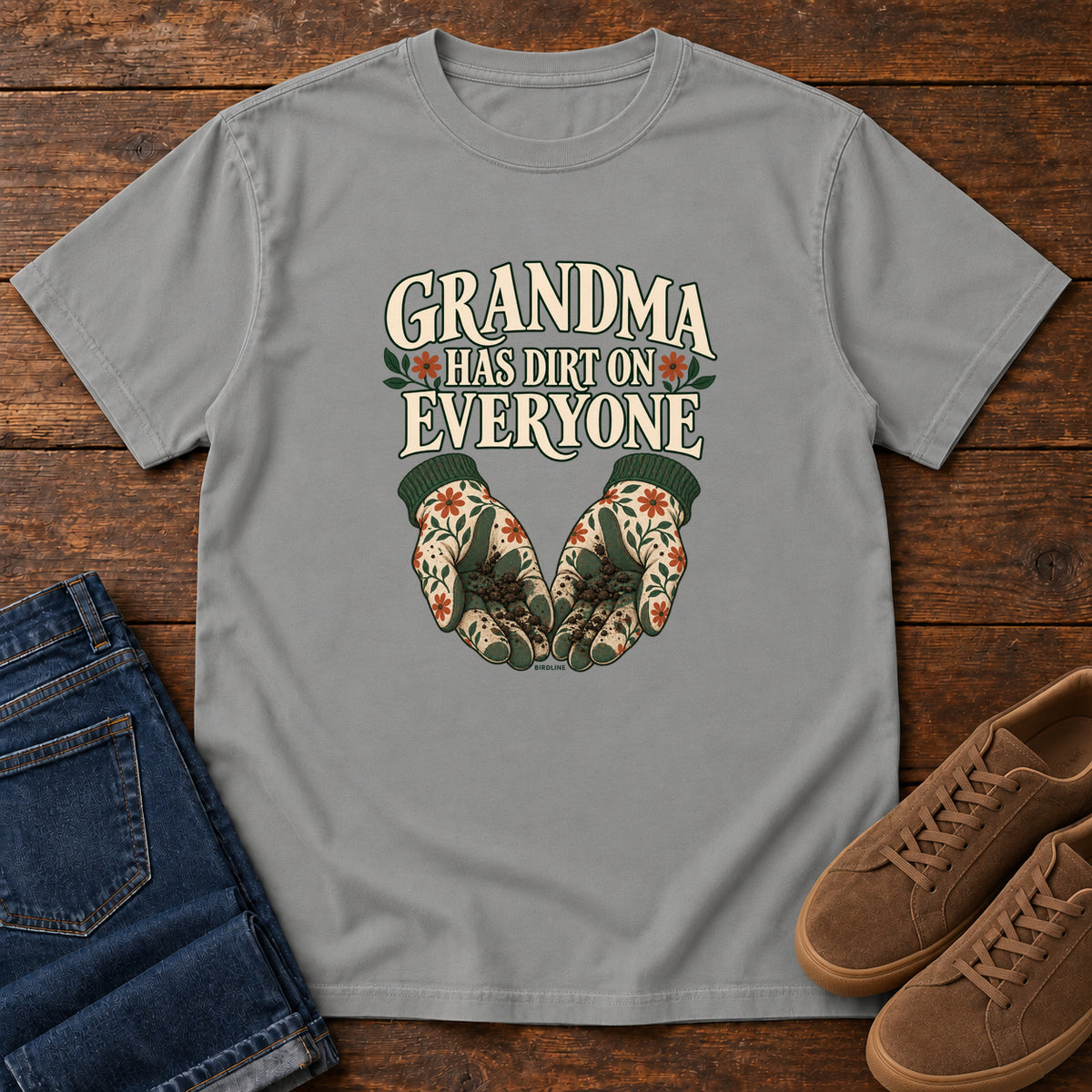 Grandma Has Dirt on Everyone T-Shirt in Grey, front laid flat on wood, featuring floral gardening gloves cupping soil beneath large cream slogan lettering.