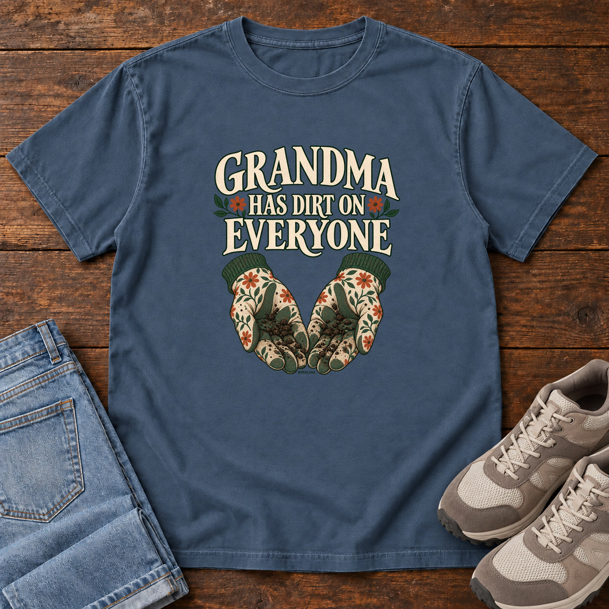 Grandma Has Dirt on Everyone T-Shirt in Midnight, front laid flat on wood, featuring floral gardening gloves cupping soil beneath large cream slogan lettering.